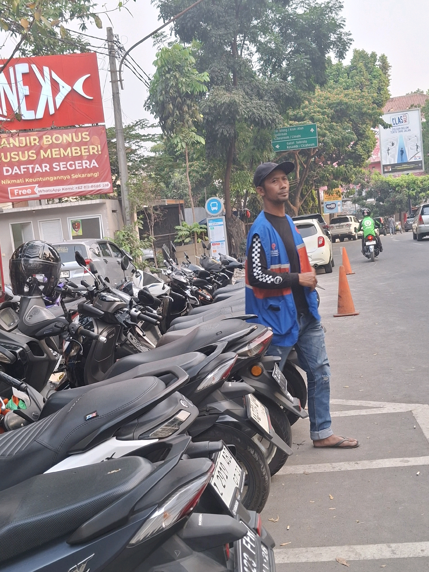 The image depicts a street scene in Bandung City, Indonesia, likely in a commercial or parking area. In the foreground, a man wearing a black baseball cap, a blue vest over a long-sleeved shirt with a checkered pattern on the sleeves, and ripped blue jeans stands next to a row of parked motorcycles. He is looking to the right with a neutral expression. His right hand is visible, holding what appears to be a small tool or object. Two orange traffic cones are placed on the asphalt road behind him.
A large red billboard with white lettering, partially obscured by trees, is visible in the background to the left. A smaller banner below it reads "ANJIR BONUS KHUSUS MEMBER! DAFTAR SEGERA Nikmati Keuntungannya Sekarang!" and has contact information. Further in the background, there are more parked motorcycles and some cars. A white car is parked on the right side of the road. A man on a green motorcycle is riding away from the camera in the background. A directional sign with Indonesian text and arrows is mounted on a pole. Other elements of the urban environment, such as power lines and trees, are also present. The overall atmosphere suggests a busy daytime scene in an Indonesian city.