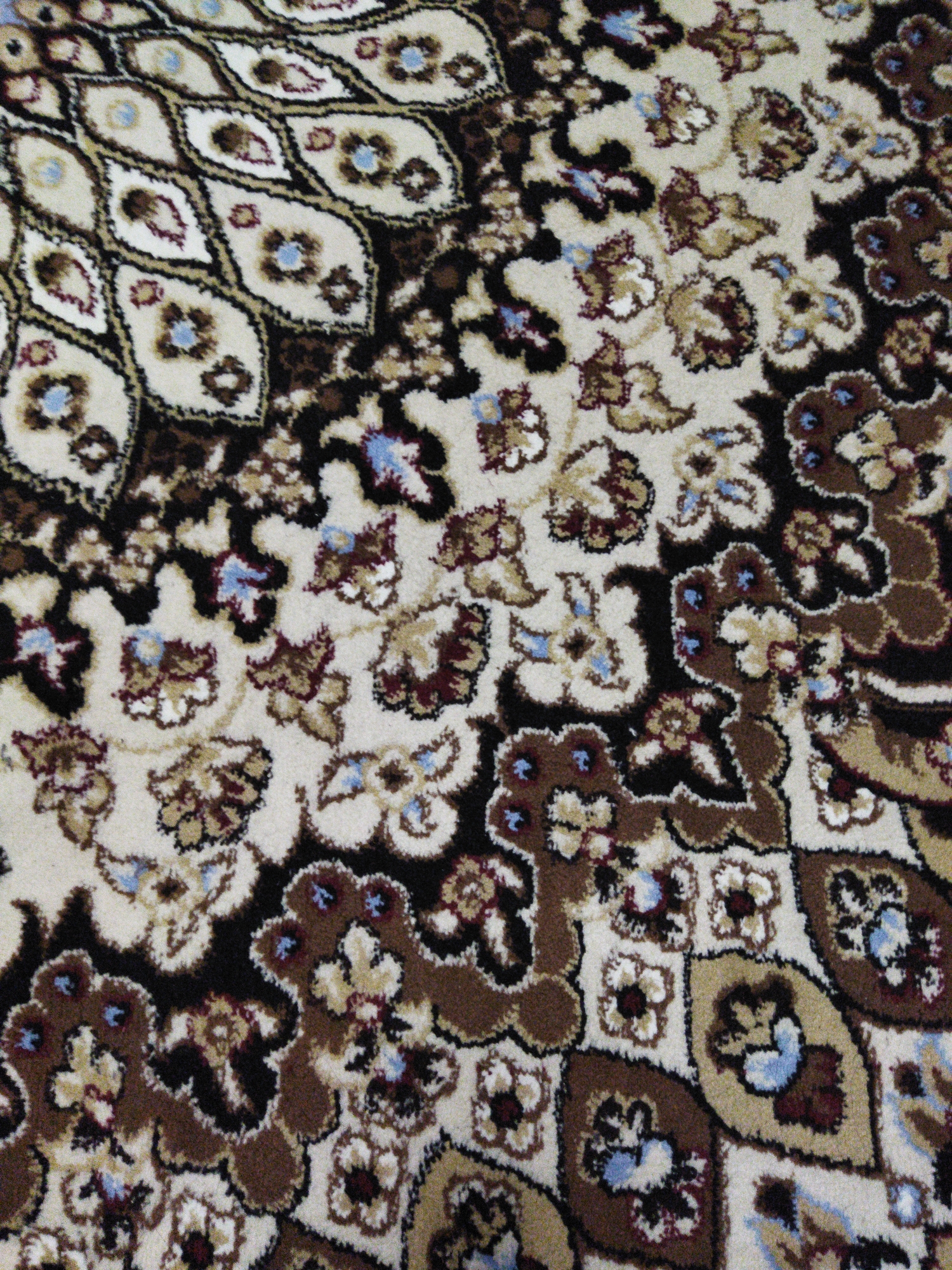 This is a close-up, overhead shot of a rug with a detailed Persian-inspired pattern. The rug features a cream-colored base with intricate designs in shades of brown, black, white, and small accents of light blue. The patterns include floral motifs, paisleys, and geometric shapes, all rendered in a textured, tufted style characteristic of woven carpets. The lighting is even, suggesting an indoor setting with ambient light, possibly from a ceiling fixture or natural light filtering in. There are no people, actions, or environmental elements beyond the rug itself visible in the frame. The image is devoid of any context relating to a specific city or geographical location, including Novopokrovka, Kyrgyzstan.