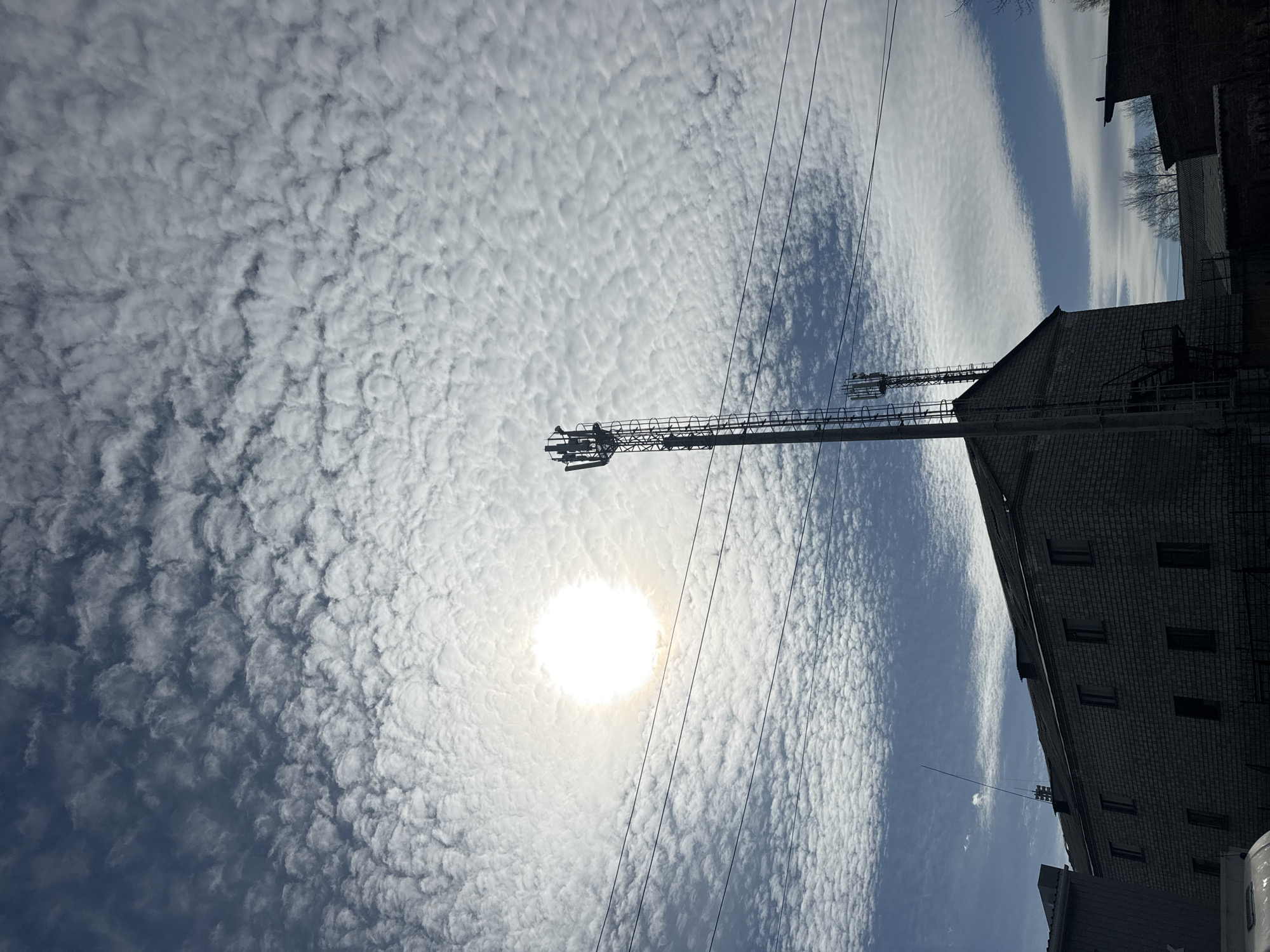 The image shows a low-angle shot of a sky filled with altocumulus clouds, taken from a street in an urban environment. In the foreground, a cellular tower with antennas is visible, obscuring part of the sky. Power lines run across the scene. The sun is visible through the clouds, creating a bright spot in the image. To the right, a tall brick building with multiple windows is present. A small part of a truck is visible in the lower-right corner. The location is possibly in the city of Krasnoyarsk, Russia, but no elements are present in the image to confirm this definitively.