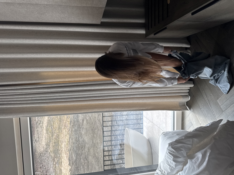 Inside a modern room, likely a hotel or apartment, in Schladming, Austria, an individual with long brown hair, wearing a white long-sleeved shirt and white knee-high socks, is depicted bending over, in the process of pulling up or taking off light blue jeans. The floor is covered with a herringbone wood pattern. To the left, a portion of a white, rumpled bed is visible. Behind the individual, light beige curtains are drawn, partially revealing a large window or sliding door. Beyond this opening is an outdoor tiled balcony featuring a freestanding white bathtub and a dark metal railing. The exterior view shows sparse, brownish-yellow ground, possibly natural terrain. To the right, a dark brown low cabinet or luggage rack is present.