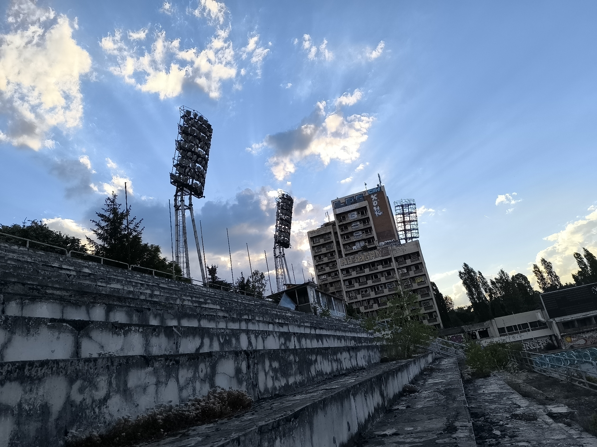 This media file depicts an abandoned stadium under a partly cloudy sky, likely at dusk. The scene is dominated by weathered concrete stadium seating, overgrown with sparse vegetation. In the background, a tall, multi-story building stands derelict, adorned with graffiti. Two large stadium floodlights are visible, one to the left and another further back, towering over the structure. The overall mood is one of decay and desolation, amplified by the fading light. The sky features dramatic crepuscular rays filtering through the clouds. While no people are visible, the setting implies past human activity, now replaced by nature's reclamation. Graffiti on the building includes the text "XOH" and "HCKC." The location is identified as Sofia, Bulgaria.