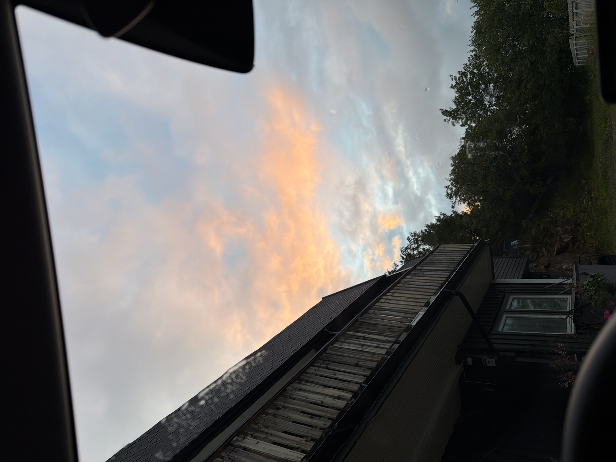 The image depicts a view of a house with a balcony and a section of a roof, against a cloudy sky with hues of pink and orange, suggesting either sunrise or sunset. The house appears to be made of dark wood siding and has a wooden railing on the balcony. There are trees visible in the background, and the overall impression is that of a residential area, possibly in a suburban or rural setting. The perspective suggests the photo was taken from inside a vehicle, possibly through a window. The location is identified as the city of Harstad, Norway.