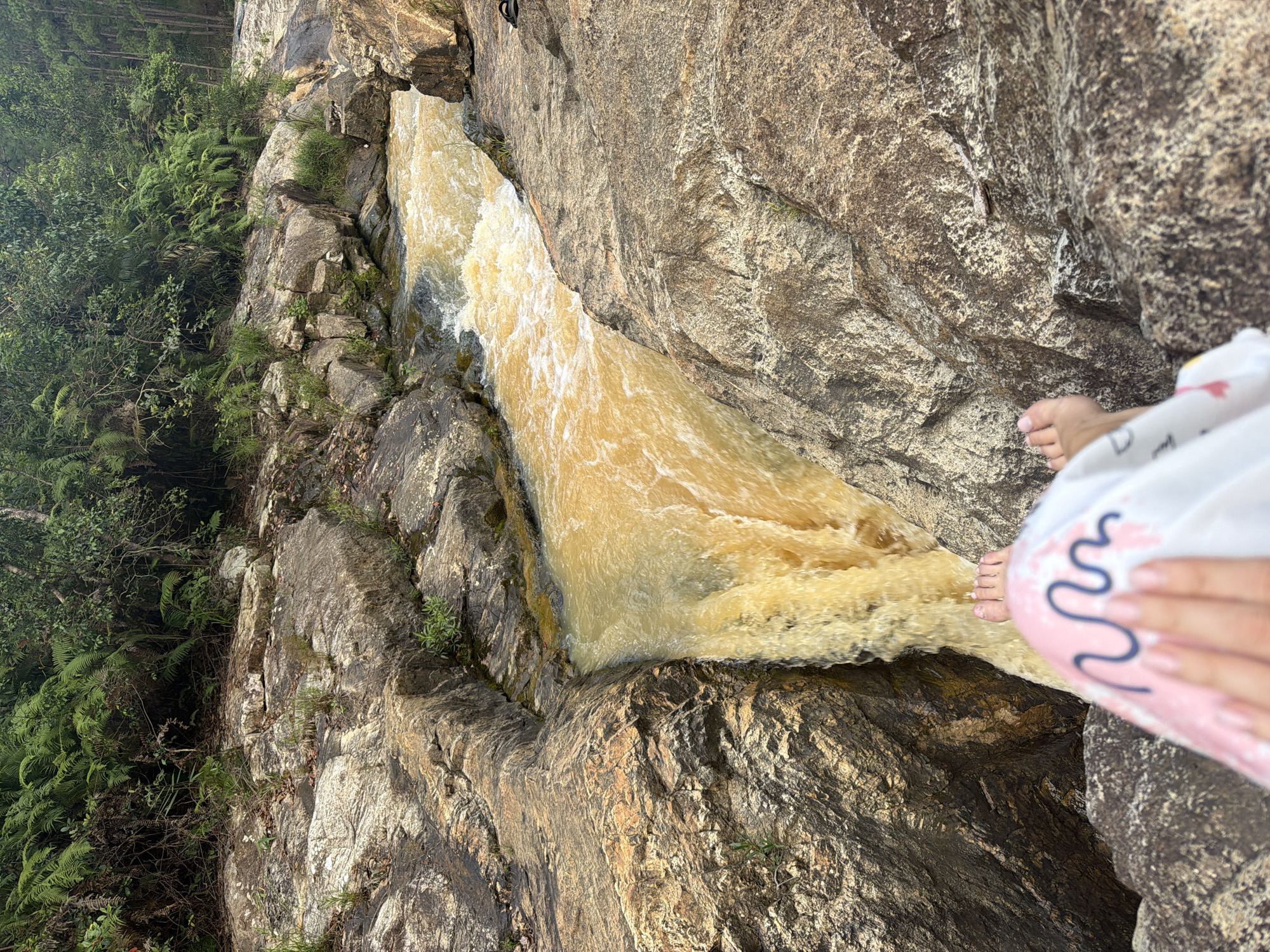 A person's bare feet, adorned with a light-colored garment patterned with pink and blue wavy lines, rest on the edge of a rocky outcrop overlooking a powerful waterfall. The water, a muddy yellow-brown, cascades forcefully down a narrow channel between rough, grey and tan rocks. Lush green ferns and trees form a dense backdrop behind the rocky terrain, suggesting a natural, possibly forested, environment. The scene appears to be a remote outdoor location, likely a natural attraction within the vicinity of Păng Tiêng 1, Vietnam.