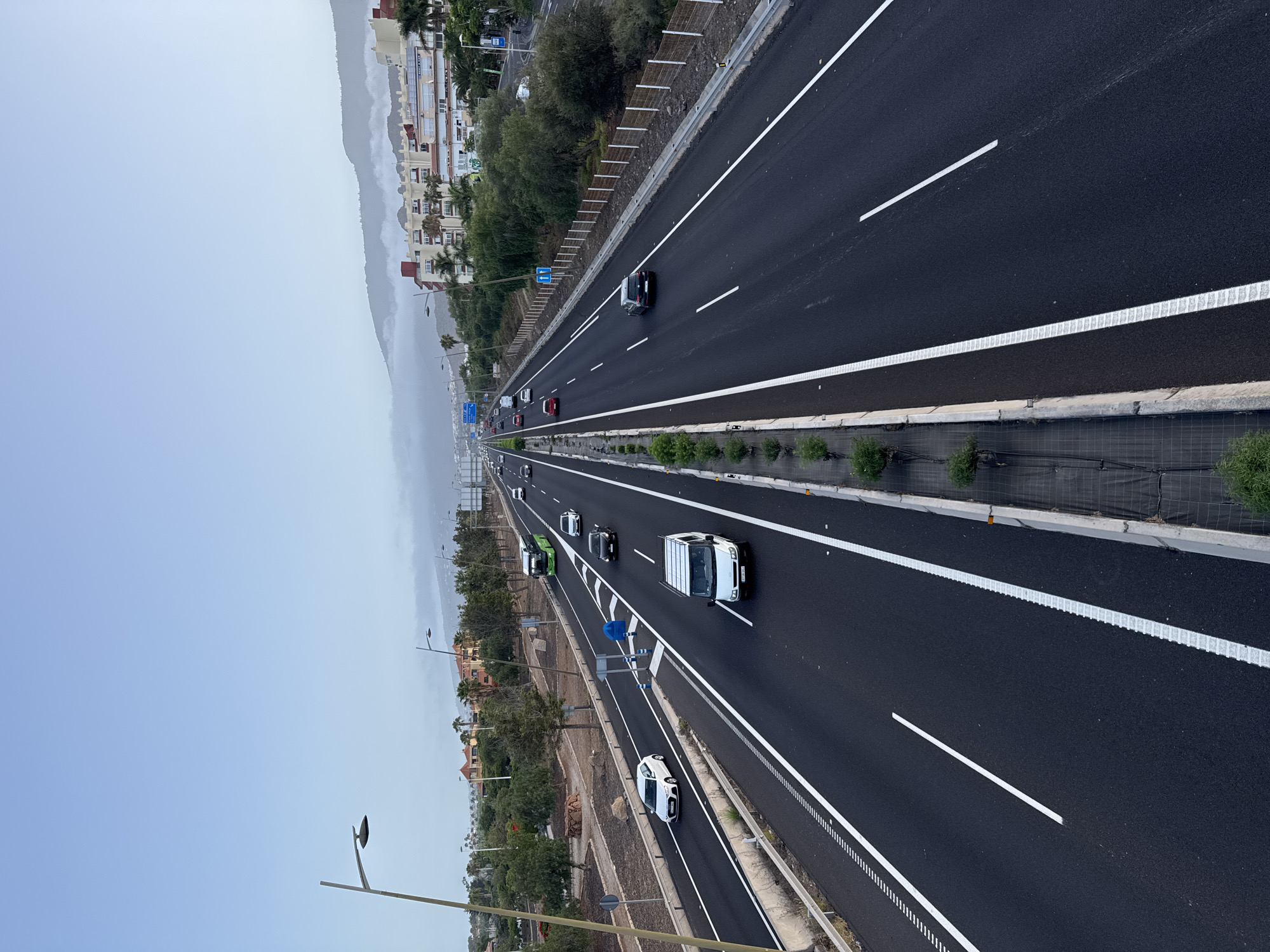 This is a medium-angle, high-contrast, full shot of a multi-lane highway in Adeje, Spain, under a hazy, overcast sky. The highway runs diagonally from the bottom center towards the top right of the frame, with a concrete median dividing opposing traffic. The asphalt is dark and wet, with white lane markings and a textured rumble strip on the edges.
On the left side of the highway, several cars are traveling in the same direction: a white hatchback, a white van, and a green bus. There are also trees, rocks, and some vegetation bordering this side of the road, with buildings and more trees visible in the distance.
On the right side of the highway, a black SUV is visible, along with several other cars further down the road. This side also features vegetation and buildings in the background. The mountains in the distance are partially obscured by fog or clouds. Streetlights are positioned along the highway on both sides. The scene depicts a typical day on a major road, with vehicles in transit and the surrounding urban and natural landscape.