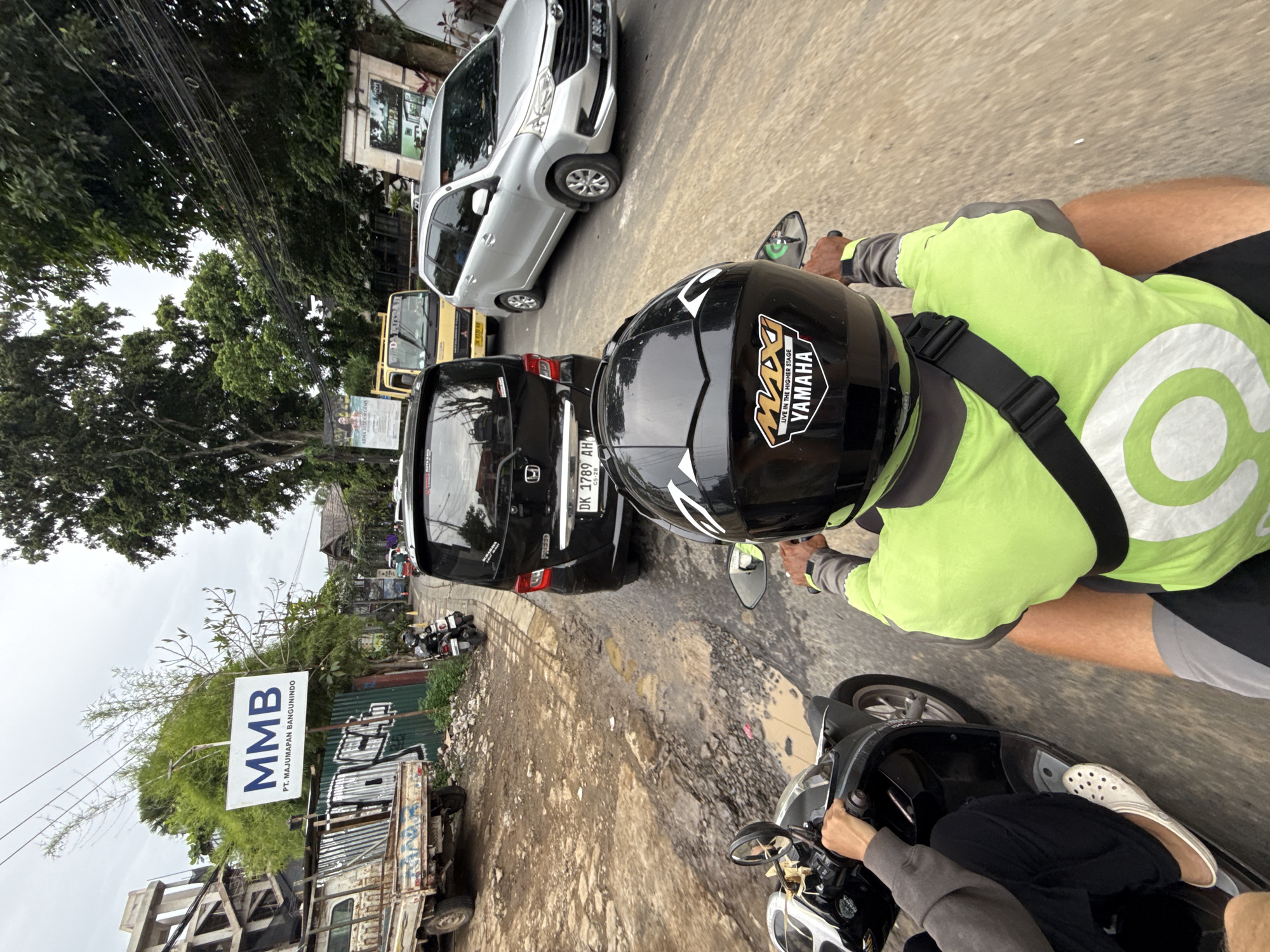 A first-person perspective captures a street scene in Ubud District, Indonesia, from the viewpoint of someone riding a scooter. The image is taken during the daytime under an overcast sky.
Ahead, another person on a scooter is visible from the back, wearing a bright neon green and grey jacket with a prominent white "Go" logo (likely indicating a Gojek driver) and a black helmet. The helmet features "MAXI YAMAHA LIVE IN THE HIGHER STAGE" stickers. The driver is wearing shorts. The person in the foreground is also on a scooter, with their hands on the handlebars and feet (wearing white clogs) visible.
The scene depicts a busy, somewhat congested road with vehicles. Directly in front of the scooters is a black Honda Freed car with license plate "DK 1789 AH" and "05-28" visible. Behind it, a yellow truck or bus is partially seen, followed by a silver Toyota Avanza-like car with license plate "DK 1904 DK." On the left side of the road, an older pick-up truck with a wooden flatbed is parked near a green corrugated fence.
The road surface appears to be in poor condition, featuring numerous large potholes and muddy, uneven patches. Utility lines crisscross overhead. Lush green trees and foliage line both sides of the road, and some buildings are visible in the background, including one with a traditional roof style. On the left, a large white sign is prominently displayed, reading "MMB PT. MAJUMAPAN BANGUNINDO." The overall impression is of a bustling, somewhat rustic urban or semi-urban environment.