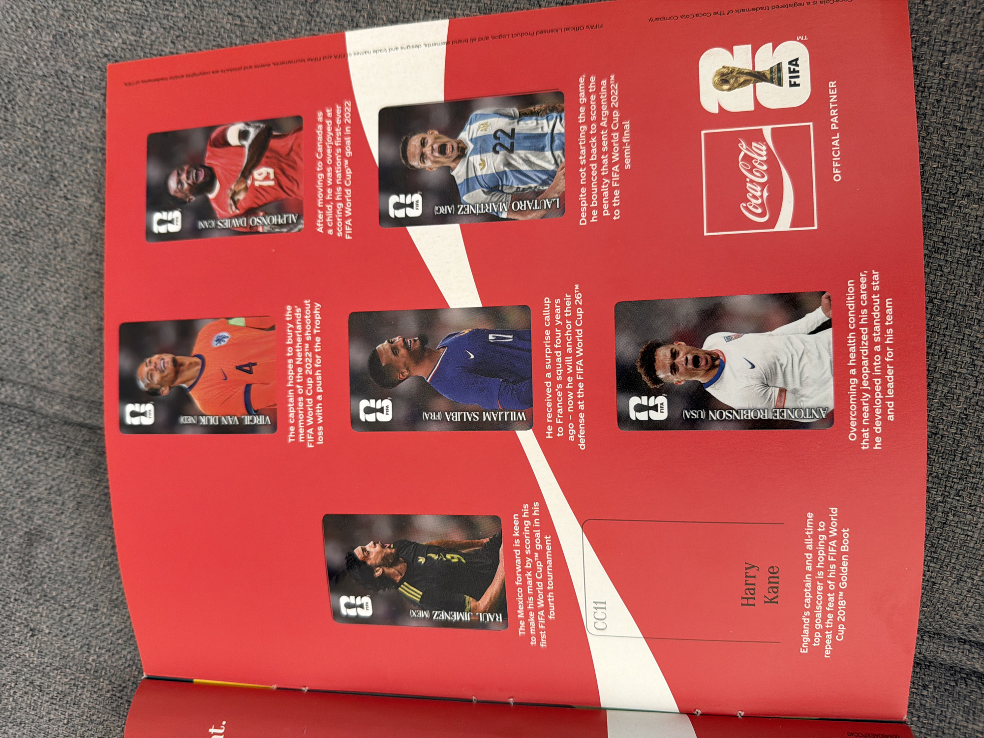 The image shows an open book with a red cover, laid flat on a textured gray surface, likely a carpet or fabric. The left page has a white section with the text "CC11" in black, followed by "Harry Kane" in black text, and then a paragraph describing him. The right page features six portrait-style cards of soccer players, each with a caption and descriptive text.
The players featured are:
* Virgil van Dijk (NED)
* Alphonso Davies (CAN)
* Raul Jimenez (MEX)
* William Saliba (FRA)
* Lautaro Martinez (ARG)
* Antonee Robinson (USA)
Below the players' cards, there are Coca-Cola and FIFA logos, with the text "OFFICIAL PARTNER".
The image depicts a spread from what appears to be a collector's book or magazine related to the FIFA World Cup, possibly a commemorative item from Coca-Cola. There is no indication of the city of Zlatitrap, Bulgaria, within the image content.