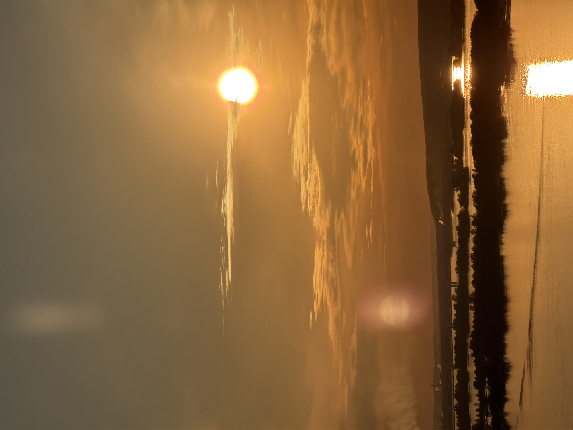 The image shows a golden sunset over a body of water, potentially a lake or wide river, with the shoreline visible on the right side of the frame. The sun is a bright, circular orb, slightly obscured by thin clouds. Its reflection shimmers on the water's surface. The overall color palette is warm, dominated by shades of orange, gold, and brown.
Without additional visual cues, it is not possible to confirm this scene as located in Innopolis, Russia.
