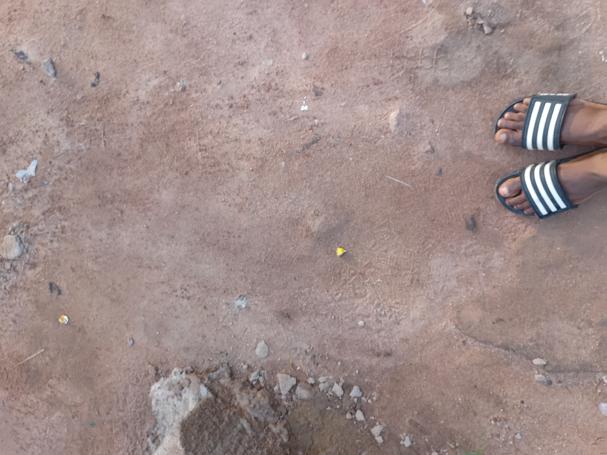 This image shows a close-up, aerial view of a person's feet wearing black and white striped slides, standing on a dirt ground. The ground appears to be a mixture of reddish-brown soil, small pebbles, and some scattered debris, including a yellow piece of paper or wrapper. To the left of the feet, there is a rough, greyish-brown concrete-like structure and some smaller rocks. The overall environment suggests an outdoor, possibly undeveloped or construction area. The scene is located in Jauro Yinu, Nigeria.