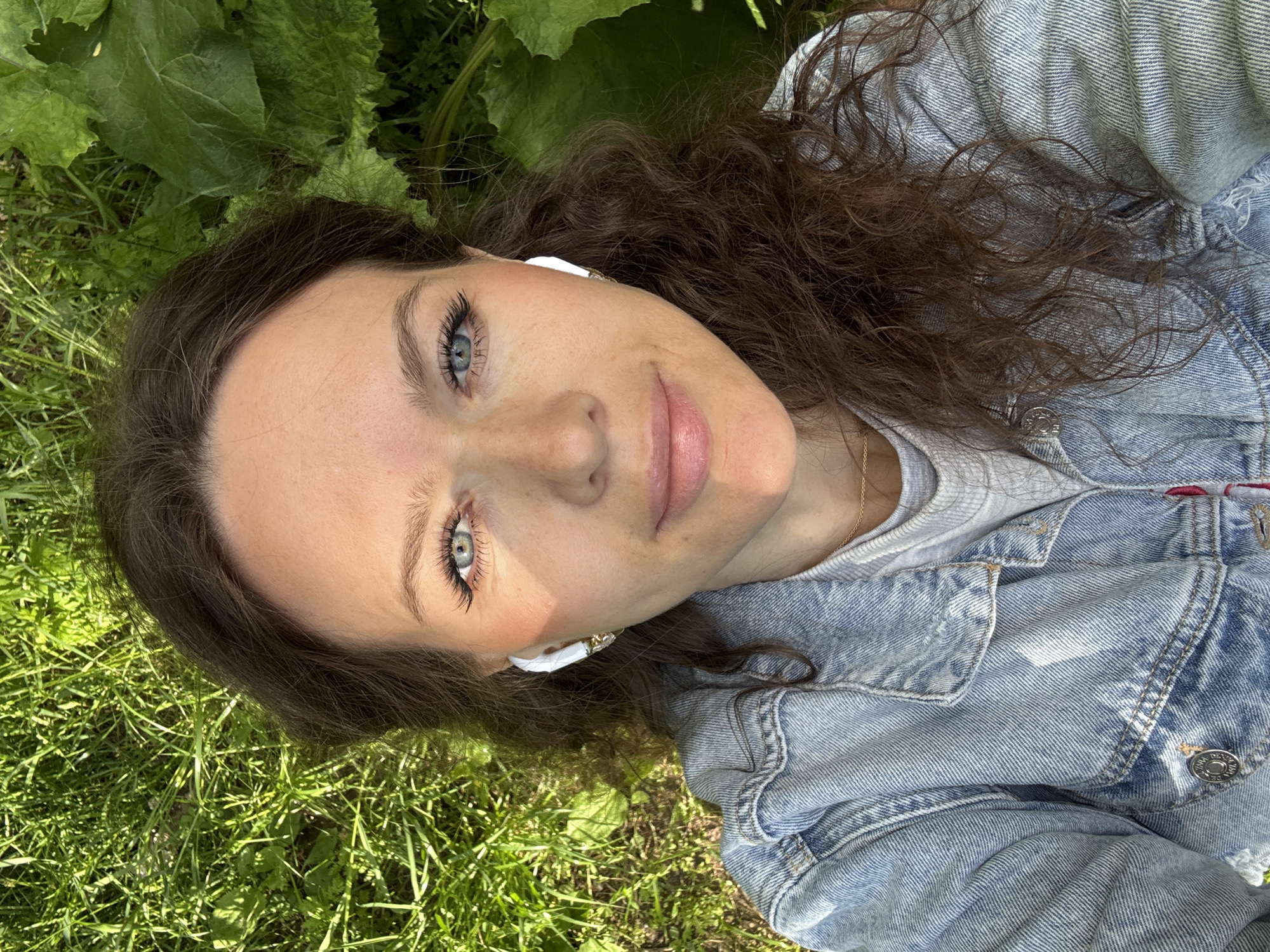 A young woman with long, curly brown hair and blue eyes is lying on a patch of green grass. She is wearing white earbuds and a light blue denim jacket over a grey shirt. Her head is slightly tilted towards the camera. Some green leaves are visible behind her head. The location is unknown.