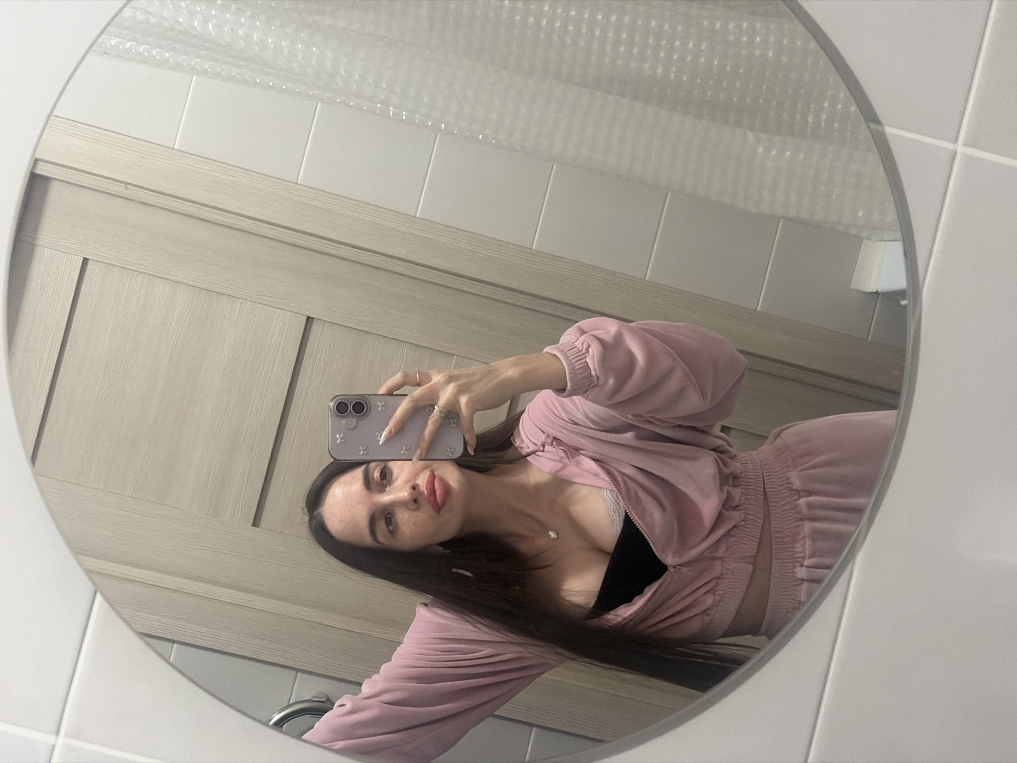 A young woman with long brown hair and light skin takes a selfie in a round mirror. She's wearing a pink velour tracksuit with a black top underneath and a silver necklace. Her left hand holds an iPhone, and she has rings on her fingers. The background reflected in the mirror includes a light wood cabinet, white tiled walls, and bubble wrap covering a portion of the upper frame. The scene appears to be inside a bathroom, possibly in Saint Petersburg, Russia, given the European-style decor.