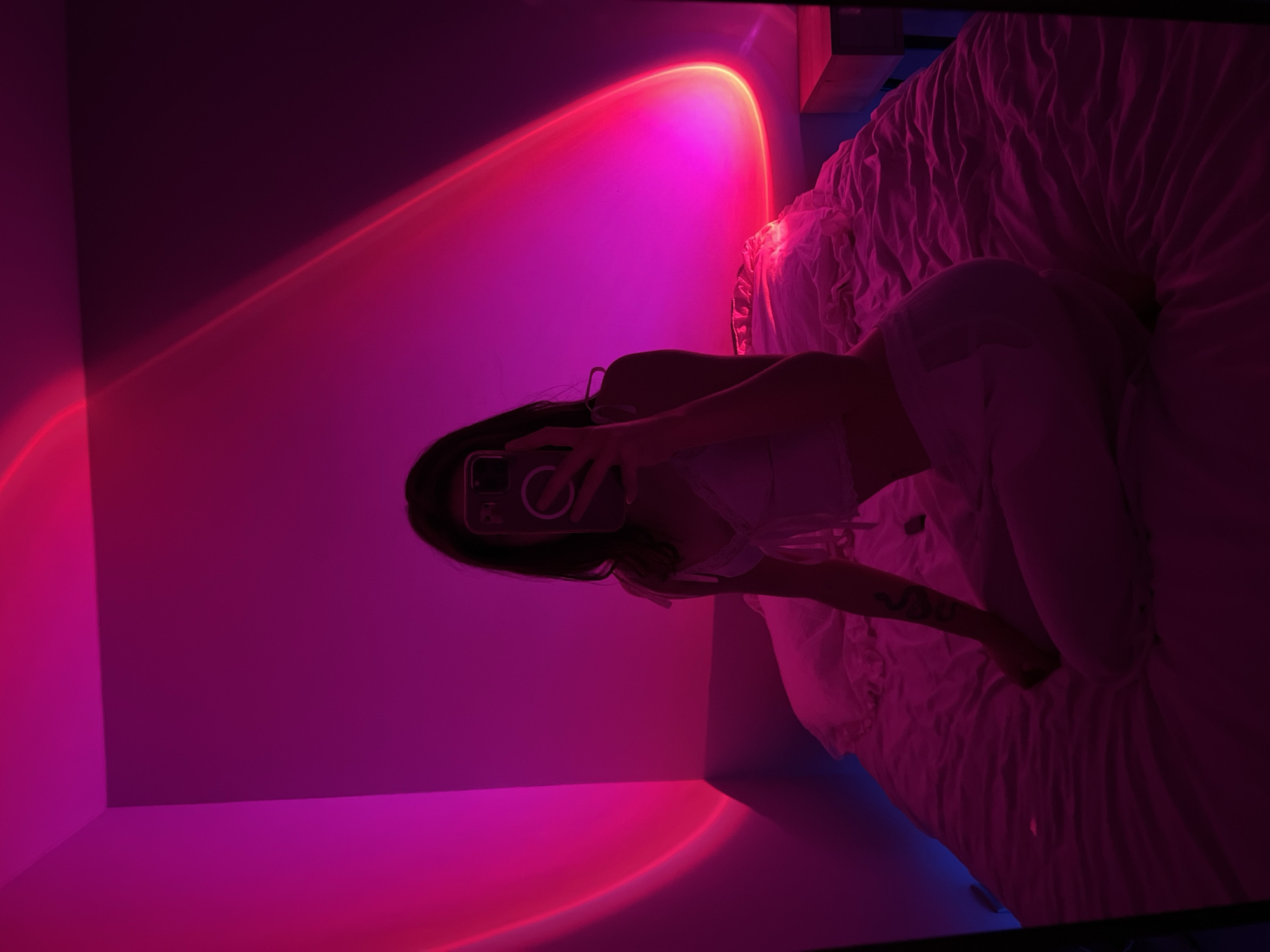 A person is taking a mirror selfie in a bedroom. The room is dimly lit with pink and purple ambient lighting. The person is sitting on a bed, holding a phone up to take the picture. The bed is covered with white sheets and pillows. The walls of the room are also illuminated by the pink light, creating a glowing effect. The overall scene is moody and intimate. The location of the image is not discernible from the provided image and crops.