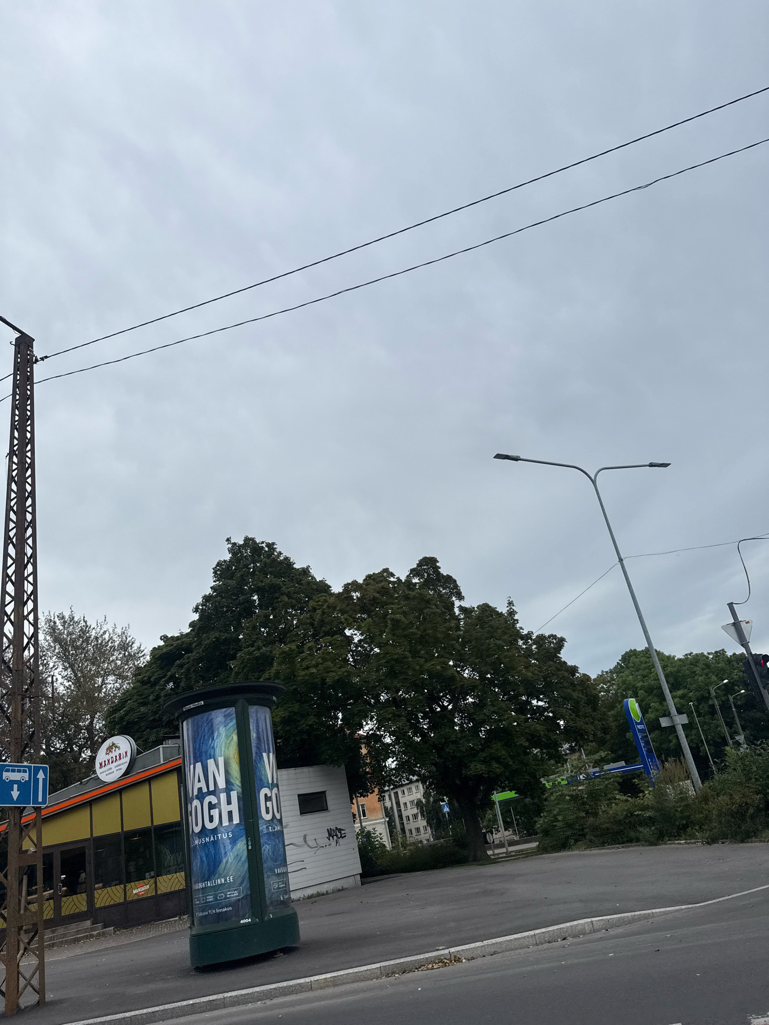 An exterior, eye-level, medium shot captures an overcast day in Tallinn, Estonia. A large, cylindrical advertisement column stands prominently in the foreground, featuring a vibrant display for a "Van Gogh" exhibition, with text in English and Estonian. The column is positioned on a paved sidewalk adjacent to a roadway. To the left of the column, a building with a yellow facade and an orange awning is visible, bearing a sign that reads "MANDARIN" with a stylized sun logo. A dark pole with a blue and white bus stop sign depicting a bus and an arrow pointing left is also situated on the left side. Behind the advertisement column, a dense canopy of dark green trees forms a backdrop. In the background, a modern street lamp with two lights extends upwards, and a blue and green vertical sign, possibly for a gas station, is partially visible among the trees. A tall, lattice-like metal structure, likely a pylon, stands to the far left of the frame. The sky is uniformly gray and cloudy.