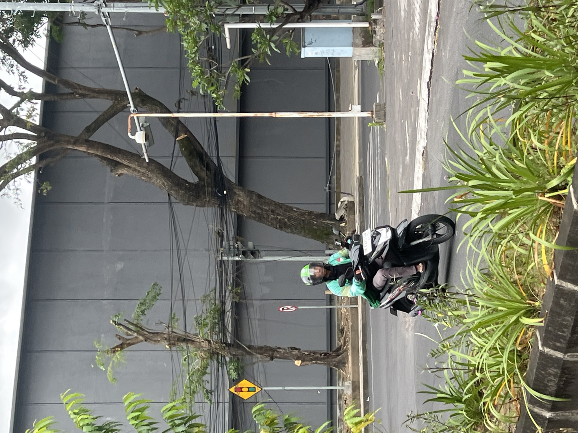 The image shows a person on a motorcycle on a city street, likely in Kuta, Indonesia. The person is wearing a helmet and a green jacket. They are riding on the right side of the road. The background includes a tall gray building, trees, and various utility poles with wires. Street signs are visible. There is some greenery along the side of the road.