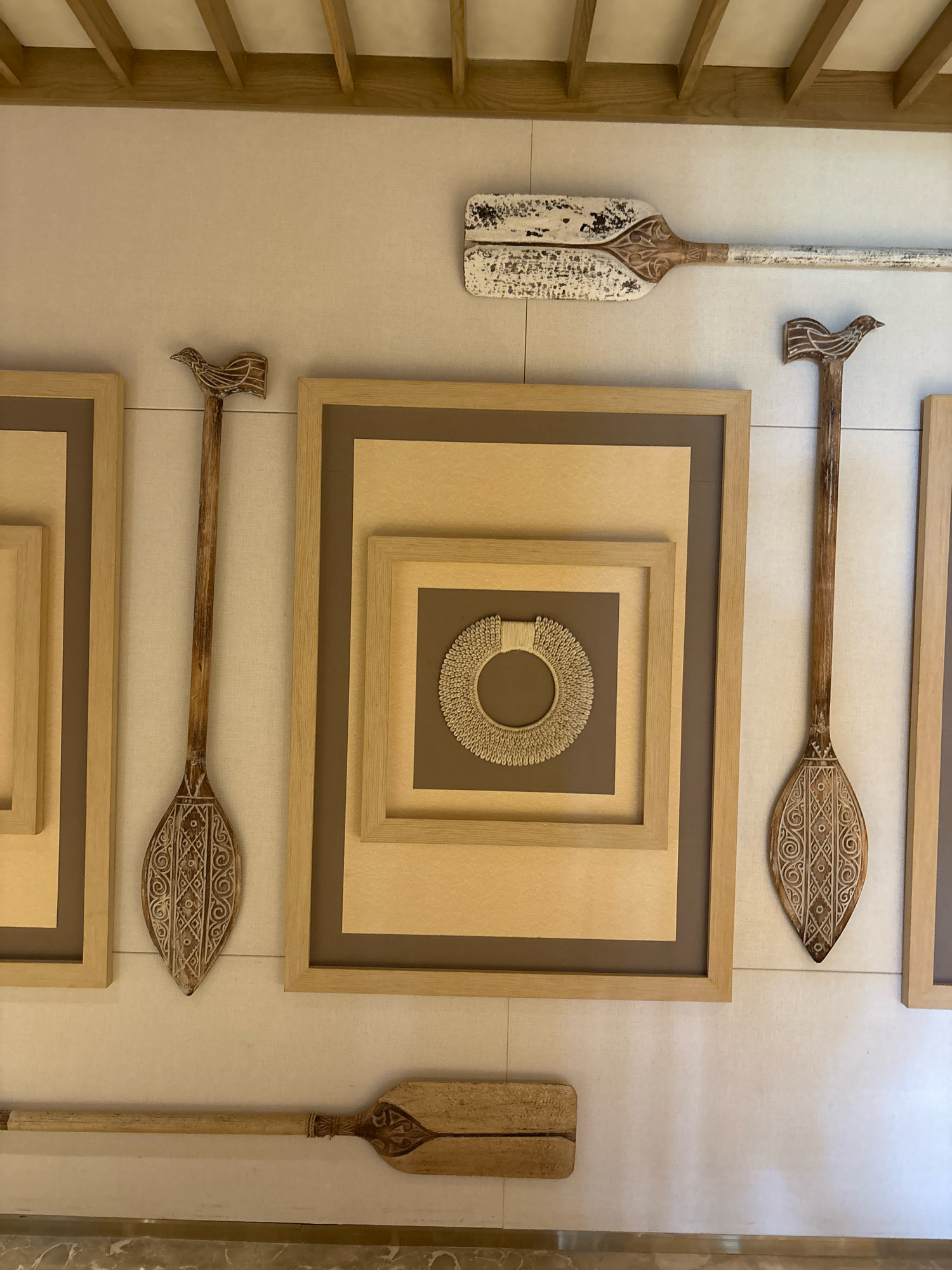 The image shows a wall decorated with wooden paddles and framed art pieces. In the center, there is a large framed artwork with multiple nested square frames, showcasing a circular woven piece. To the left and right of this central piece are two vertical wooden paddles with intricate carvings on their blades, one of which has a bird-like handle. Above the central artwork, a weathered white and wood paddle is mounted horizontally. Below, another wooden paddle is positioned horizontally. The wall is a light, textured color, and the ceiling features wooden beams. The floor visible at the bottom is marble. This scene is located in Vagator, India, specifically at a restaurant called Titlie.