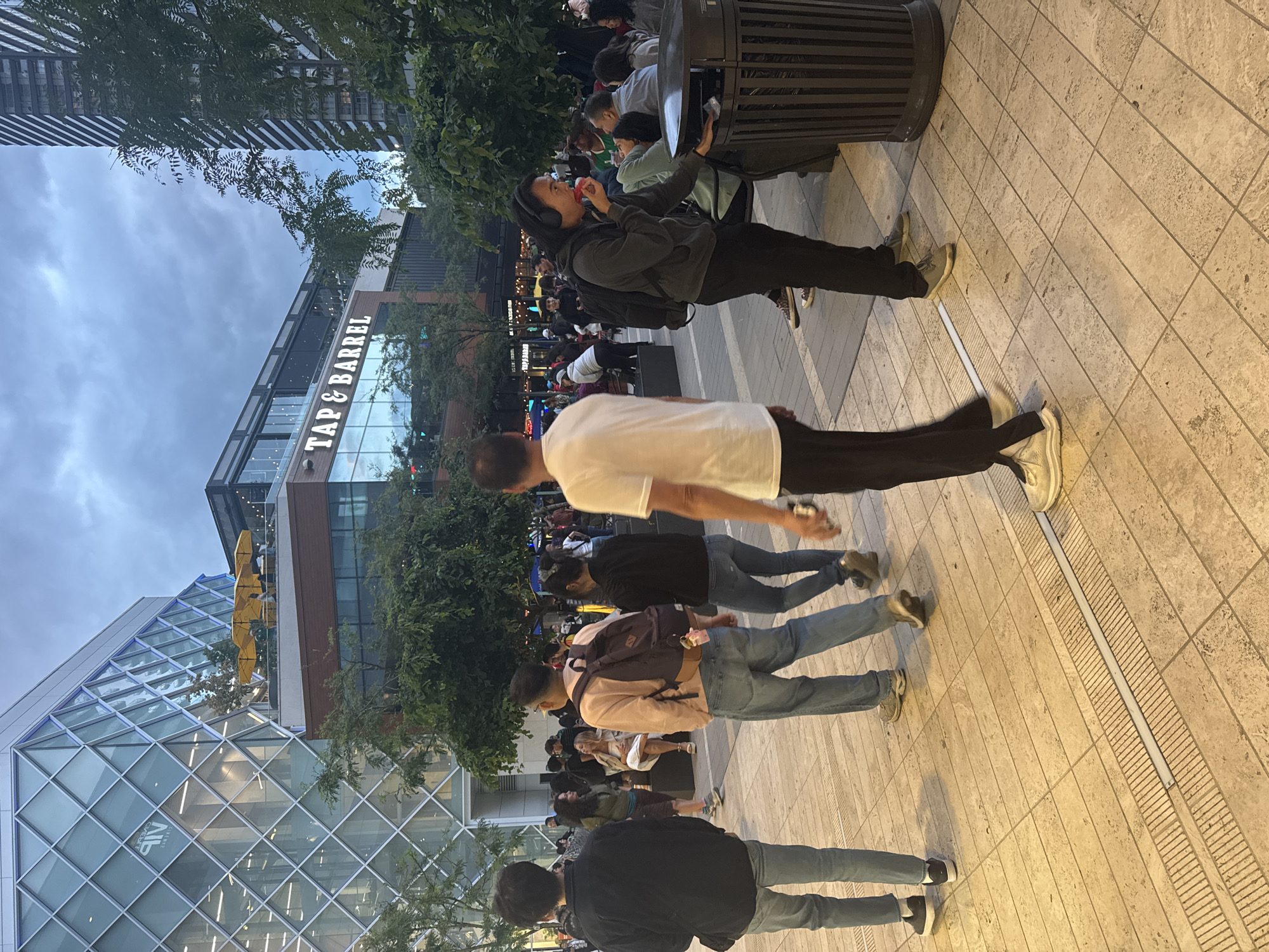 In an outdoor urban plaza in Burnaby, Canada, a group of people are gathered in the late afternoon or early evening. The architecture features a modern building with a diamond-patterned glass facade, and another building displaying a "TAP & BARREL" sign. The ground is paved with light-colored tiles.
Several individuals are visible. In the foreground, a person wearing a black jacket and light-colored jeans walks away from the camera. To their right, another person in a light pink sweatshirt and blue jeans, carrying a backpack, is also walking away. Further to the right, a person in a white t-shirt and black pants is seen from behind. In the middle ground, a person wearing a grey hoodie and black pants with headphones is eating something red while standing near a dark, cylindrical trash receptacle. Other individuals are dispersed throughout the plaza, some sitting and some standing, engaged in conversations or simply present in the area. Trees with green foliage are scattered throughout the scene, and the sky is overcast, suggesting twilight or a cloudy day.