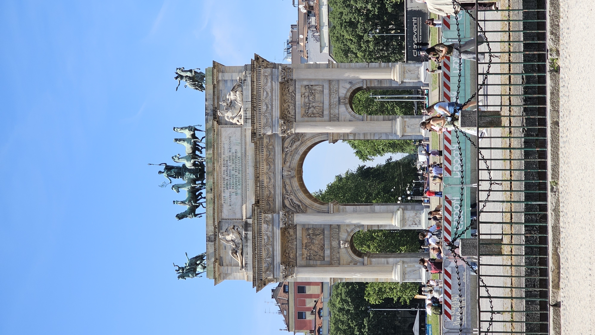 The image shows the Arco della Pace in Milan, Italy, viewed from ground level. The marble arch is adorned with sculptures and carvings, including a bronze statue atop depicting a chariot pulled by horses. In the background are trees, other buildings and a clear blue sky. A fence runs across the foreground, with some people near the barriers. Signs or banners are affixed to the fence. The archway provides a portal view of the park beyond.
