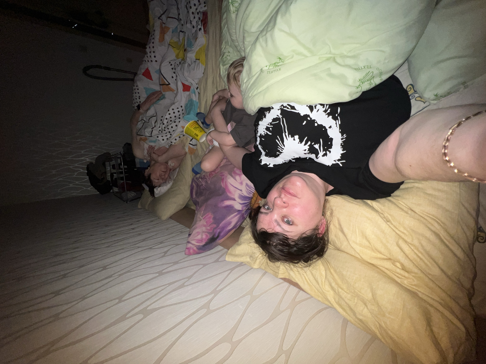 A woman is lying on a bed with a patterned cream-colored comforter and a yellow pillow. She is wearing a black t-shirt with a white design. A toddler is sitting near her, and an older person is lying under patterned bedding. A green pillow is partially covering the toddler. A bottle of lotion or sunscreen is visible near the older person. A shopping cart with miscellaneous items sits near the edge of the bed. The location is indicated as Perm, Russia. The photograph appears to be taken in a residential setting.