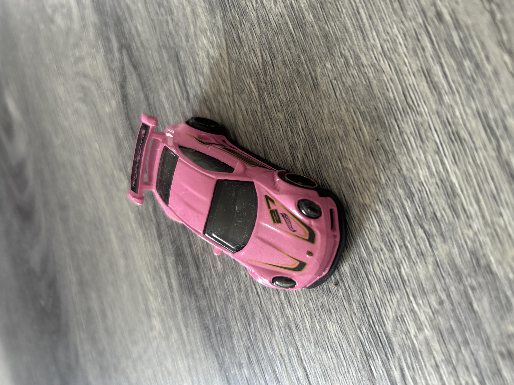 A high-angle, close-up shot reveals a pink toy car, likely a Porsche, resting on a gray, wood-grain surface. The toy car features black accents, including the wheels, windows, and decorative stripes. The car's spoiler has the name "Porsche" printed on it. The image does not depict any people, interactions, or details suggestive of Câmara de Lobos, Portugal; the scene is a generic close-up of a toy on a wooden surface.