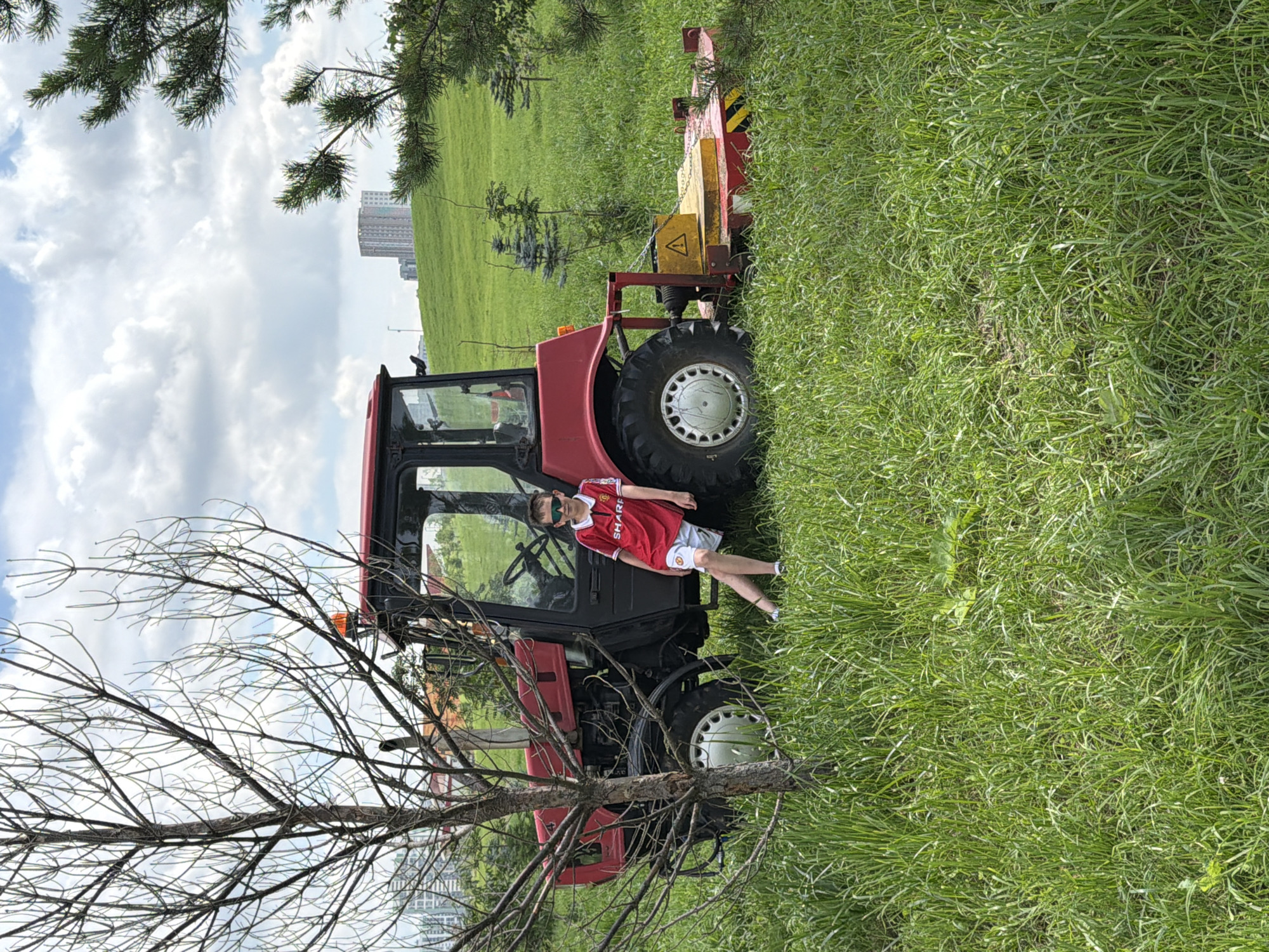 A young boy wearing a red football jersey and white shorts with sunglasses leans against a red tractor in a grassy field on a cloudy day. The tractor is parked next to a bare tree and has a mower attachment. In the background, there are green hills with some young trees and a tall building in the distance. The scene appears to be in a park or an undeveloped area within the city of Yekaterinburg, Russia.