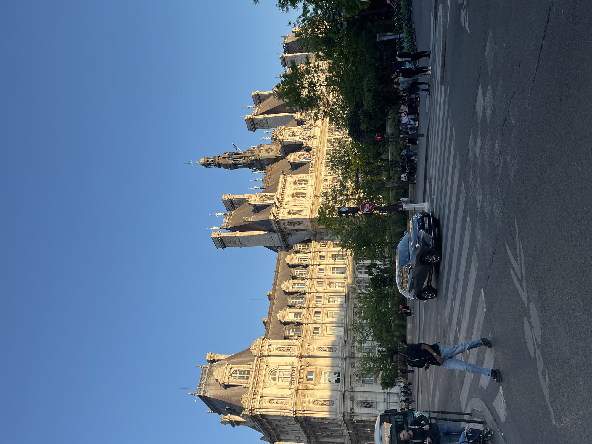 A sunlit urban scene in Paris, France, captures the grandeur of a historic building and the activity of a city street. The dominant subject is a magnificent, ornate stone building, likely the Hôtel de Ville, bathed in golden sunlight from the right, indicating late afternoon or early evening. Its facade features numerous arched and rectangular windows, dormers, intricate sculptures, and a prominent clock tower topped with a spire. Trees with lush green foliage line the street in front of the building, partially obscuring parts of its lower sections.
In the foreground, a wide asphalt street with a white-striped pedestrian crossing is visible. A dark-colored car is stopped on the crossing, facing right. Further down the street, on the left, a line of bicycles is parked. A man in a black shirt and light blue jeans is walking across the pedestrian crossing, seemingly towards the right side of the frame. He appears to be wearing earphones. On the sidewalk to the left, beneath the trees, several people are gathered, some appearing to wait at a bus stop or for a traffic signal. A red traffic light is visible above this crowd. Near the bottom left, a white bus-like vehicle is partially visible, with two individuals near it; one appears to be pushing a stroller.
A red circular "No Entry" (sens interdit) sign is visible near the street level. The sky is a clear, vibrant blue, indicating good weather. The license plate on the dark car shows "F 646 DV" and possibly more characters. Partial text "ROUTE A LA VALLÉE" is visible on the side of the white bus. The overall scene conveys a typical, bustling urban environment under clear skies, highlighting the city's architectural beauty alongside everyday life.