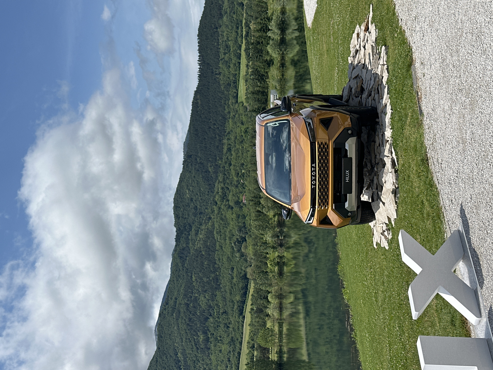 The image captures a striking golden-bronze Toyota Hilux pickup truck showcased in a picturesque outdoor setting. The truck is positioned on an arrangement of light-colored rocks, creating an off-road display in the foreground. Its front grille, clearly visible, features the prominent "TOYOTA" branding and "HILUX" model name.
Behind the vehicle, a serene body of water, likely a lake, stretches across the mid-ground, perfectly reflecting the verdant, tree-covered mountains that rise majestically in the background. The sky above is a vibrant blue, adorned with large, fluffy white clouds, indicating clear and sunny weather.
In the immediate foreground, to the right of the truck, there is a gravel path or area. This is separated from the truck's display by a strip of lush green grass. A large, modern-looking white 'X' shaped sculpture or sign is partially visible in the lower right corner, hinting at a curated display environment.
The scene depicts a bright day, likely late morning or early afternoon, given the clear sunlight. There is no specific activity taking place; the truck is stationary, suggesting a display or photoshoot. The mountainous landscape and body of water align with the specified location of Razliv, Bulgaria, creating a scenic and natural backdrop that complements the rugged aesthetic of the vehicle.