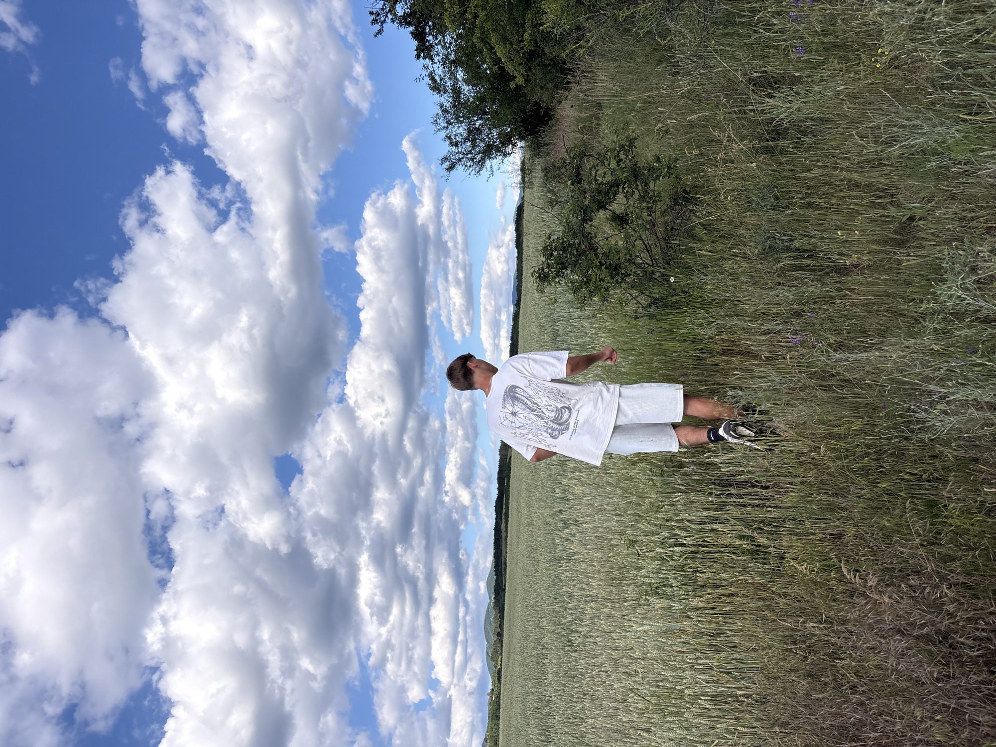 A young Caucasian male, seen from behind, walks through a field of tall, green crops under a partly cloudy blue sky. He is wearing a white t-shirt with a graphic print on the back and light-colored shorts, along with athletic shoes. The field extends towards a distant, rolling landscape of hills or mountains. To the right of the frame, there is a dense growth of trees and bushes, with tall, dry grass in the foreground. The overall impression is that of an outdoor, rural setting, possibly in the vicinity of Podgumer, Bulgaria.