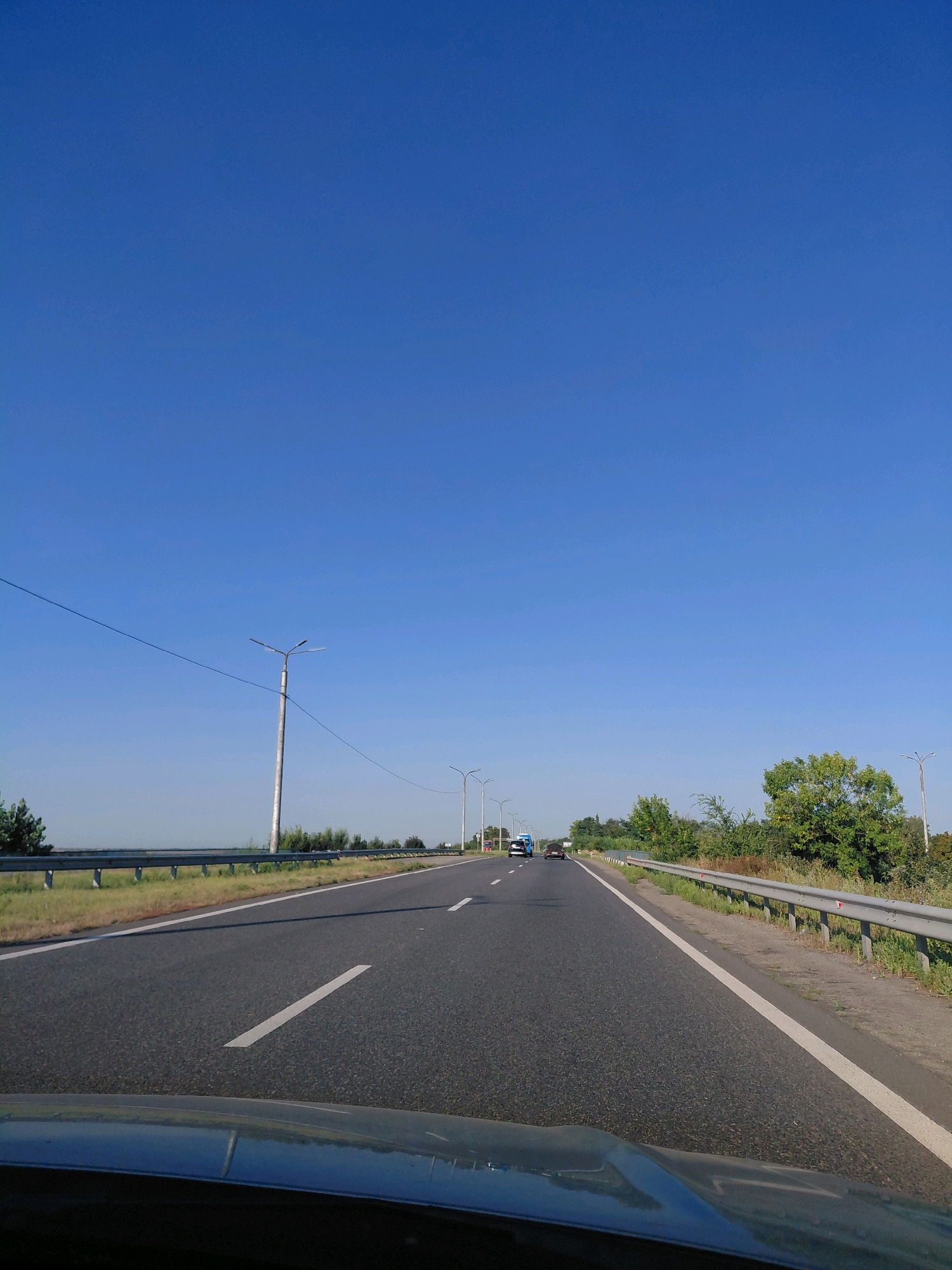 A bright, clear blue sky stretches over a highway with a guardrail on the right side. The highway has white lane markings and appears to be in good condition. Several vehicles are visible on the road, including a blue van and a black car. The surrounding landscape is green with trees and vegetation, suggesting a rural or suburban environment. The image is taken from the perspective of a vehicle driving on the highway.