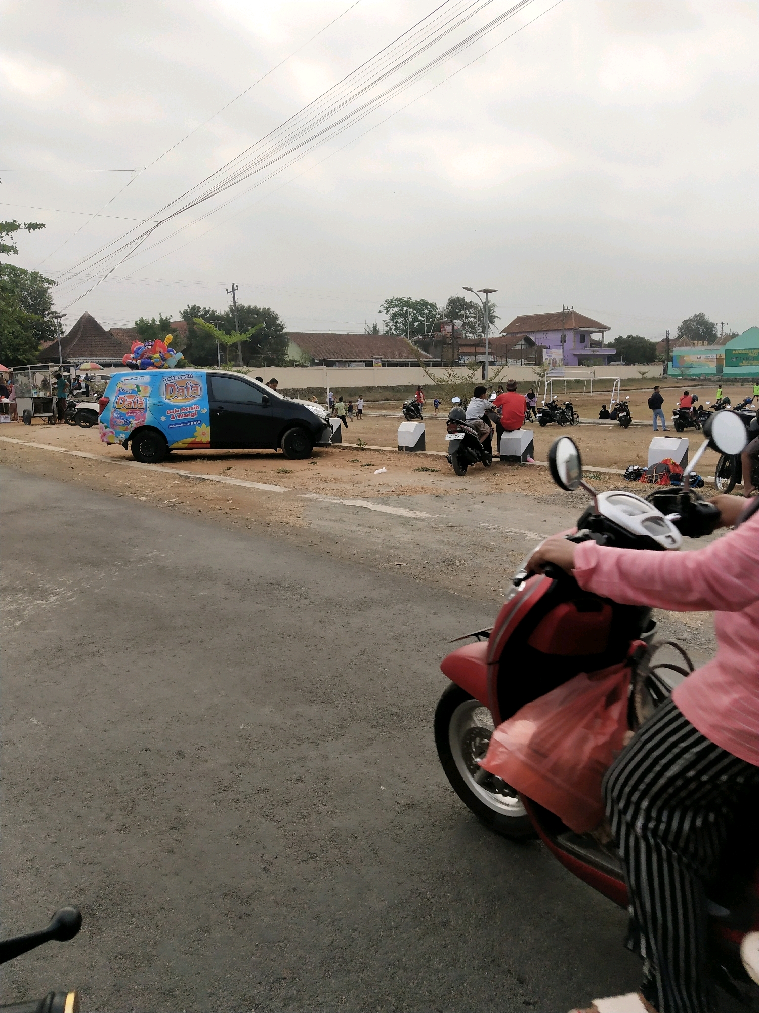 The image depicts an outdoor scene, likely a public space or road, in Desa Kedondong Kecamatan Sokaraja, Indonesia. In the foreground, the handlebars and front portion of a red scooter are visible, with a person wearing a pink shirt and striped pants on the scooter. A red plastic bag is hanging from the scooter. In the midground, a black van with colorful advertisements for "Deterjen Daia" is parked. The van is decorated with balloons on its roof. Several people are seen around the van and in the background, some on motorcycles. Other motorcycles are parked or in motion in the distance. The background features a street, buildings with reddish-brown roofs, trees, and a cloudy sky. The overall environment appears to be a street or parking area near a residential or commercial zone.