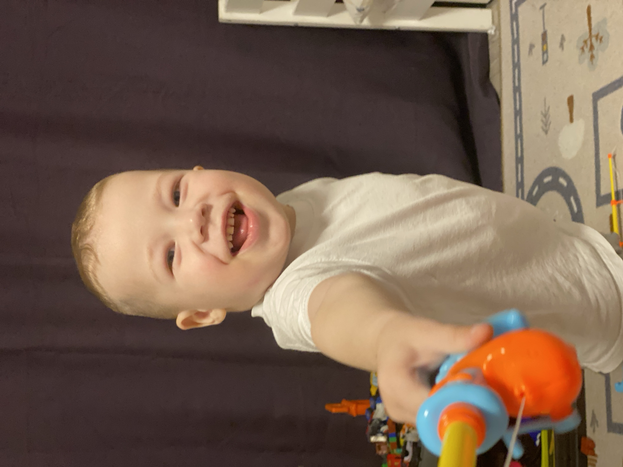 A young child wearing a white t-shirt is holding a toy, possibly an orange and blue puffer toy with a yellow handle, towards the viewer. The child is smiling broadly, revealing their teeth. The background features a dark purple curtain or wall, a white shelf, and a play mat with a road pattern. There is a collection of toys at the bottom of the frame. It is not possible to tell whether the photo was taken in Sochi, Russia.