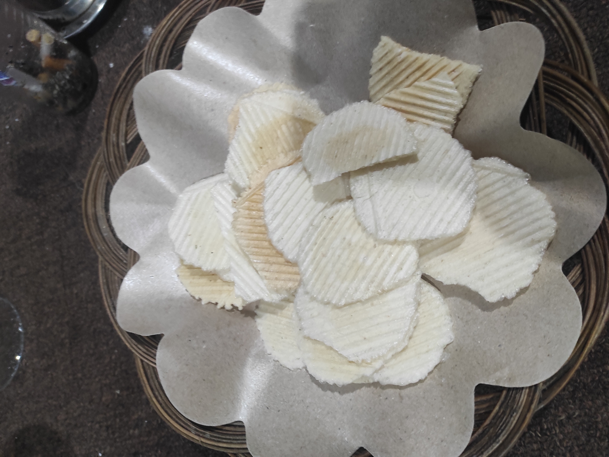 A close-up, overhead shot captures a pile of rippled, pale chips, possibly potato or cassava chips, artfully arranged on a decorative, petal-shaped paper liner. The liner itself rests within a woven basket, hinting at a rustic or traditional presentation. The chips exhibit a light, golden-brown hue in some areas, suggesting they are fried, and their ridged texture is a prominent feature. The background is a dark, textured surface, possibly a tablecloth or a table. A blurred glass and a small, clear jar containing what appear to be cigarettes are visible in the upper right corner, suggesting this scene might be set in a cafe or a dining area. The overall impression is one of a snack or appetizer being served. This image was taken in Sumedang, Indonesia.