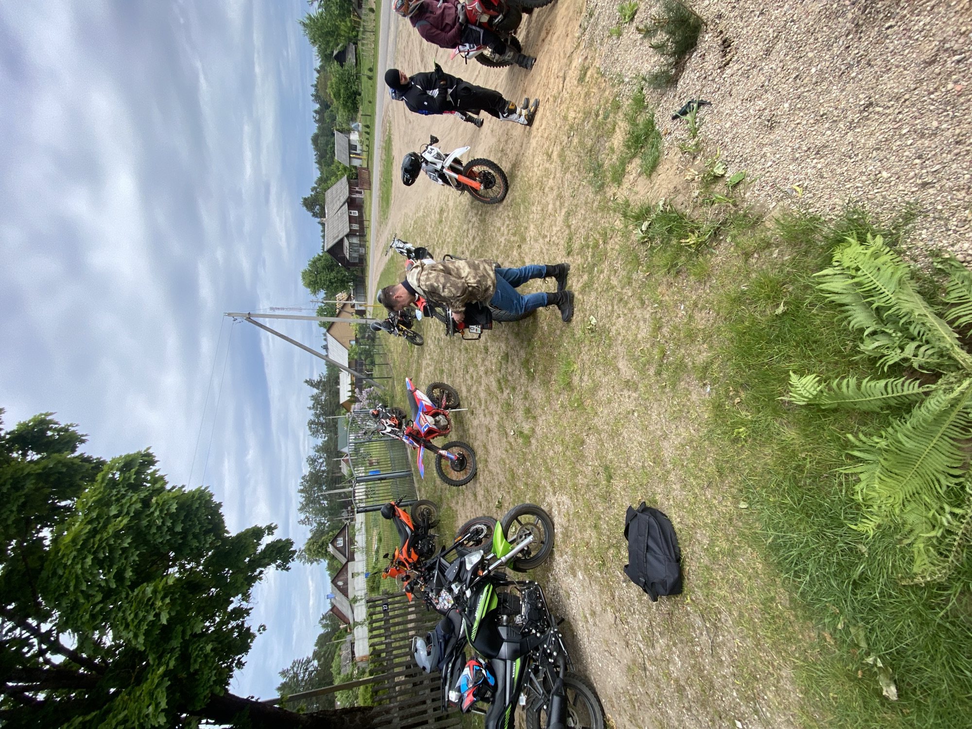 A group of people are gathered with several motorcycles in Парэцкі сельскі Савет, Belarus. One man is bent over a motorcycle, seemingly working on it. Other individuals stand near their bikes, observing. The environment consists of a grassy area with patches of gravel and dirt. There are houses in the background. A tree stands to the left.