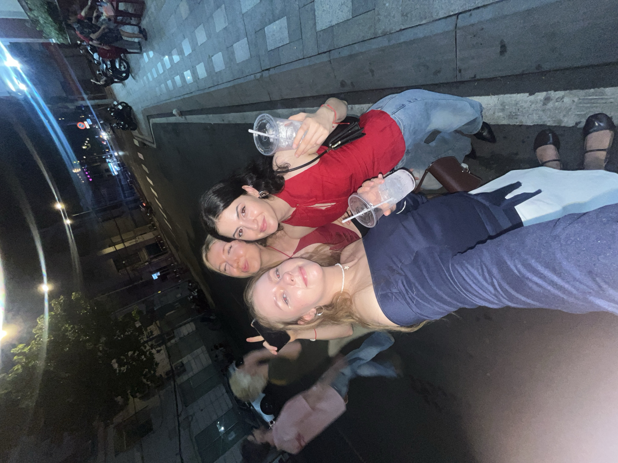 Three young women pose for a photo outdoors in Thủ Đức, Vietnam. The woman in the foreground wears a navy top, light jeans, and a white necklace. The woman in the middle wears a red top. The woman in the back wears a red top and light jeans. Each of the women holds a clear plastic cup with a straw. The background shows a city street at night with motorcycles parked along the side and streetlights illuminating buildings.