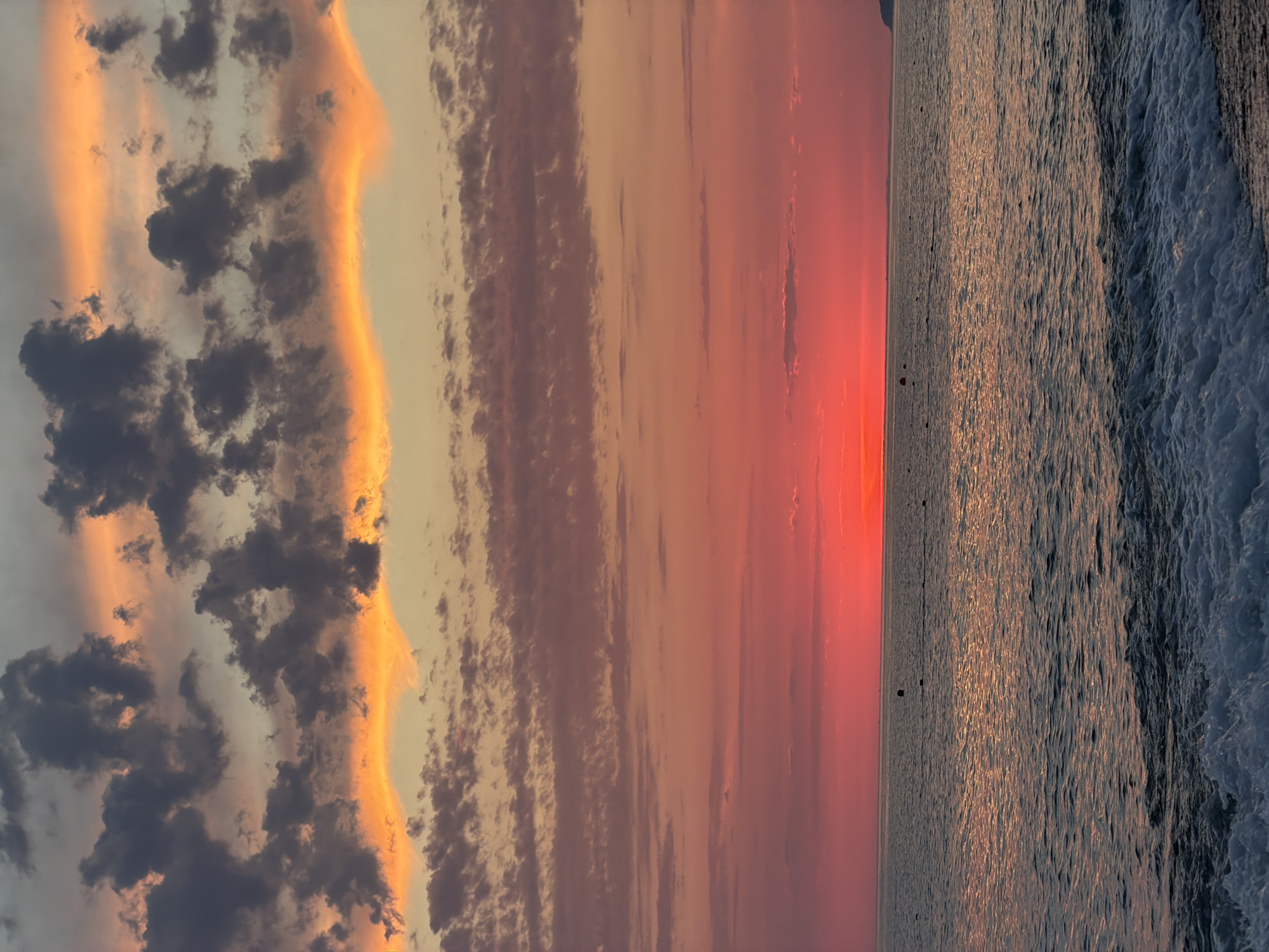 The image depicts a serene sunset over the Black Sea, likely taken from the shore in Sochi, Russia. The sky is a vibrant canvas of colors, transitioning from a bright, fiery red at the horizon to softer pinks and oranges higher up, with dramatic dark gray clouds edged in orange in the upper portion of the frame. The sea reflects the colors of the sky, with shimmering patterns of light dancing on the surface. Small buoys are visible dotting the water, indicating a possible swimming or boating area. In the foreground, gentle waves crash onto a pebble beach. The overall atmosphere is one of peaceful tranquility as day turns into night.