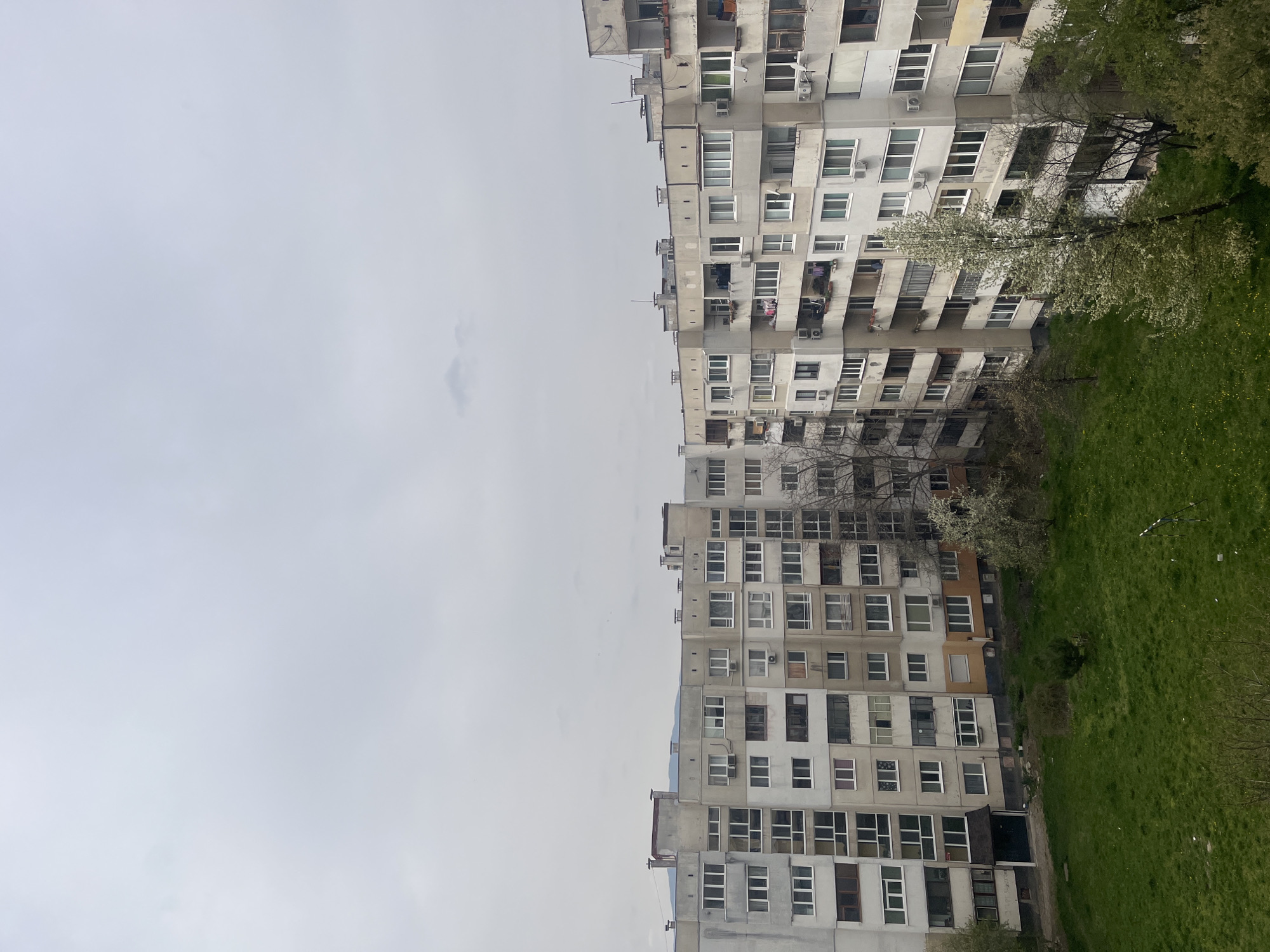The image depicts two high-rise residential buildings in Sofia, Bulgaria. The buildings, typical of Soviet-era architecture, are gray and feature numerous windows and balconies. A patch of green grass and some trees are visible to the right of the buildings. The sky is overcast and gray. No people are visible. The scene is static and devoid of activity.
