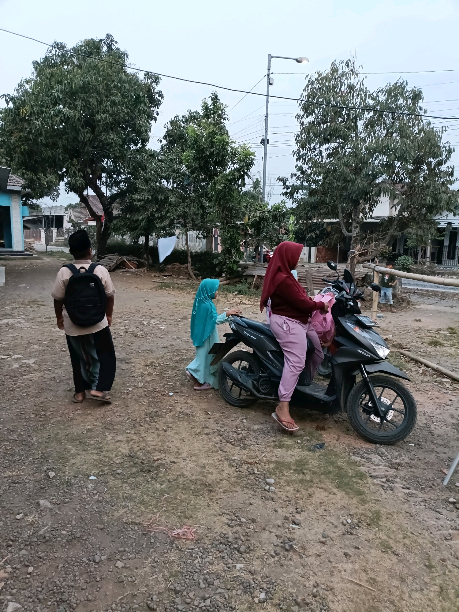 In a dusty, unpaved area in Tamanan, Indonesia, two children and an adult are near a black Honda motorcycle. A young boy stands with his back to the camera, wearing a beige shirt, dark pants, and a black backpack. To his right, a young girl in a turquoise headscarf and dress stands by the motorcycle. An adult woman wearing a maroon headscarf and light pink pants is seated on the motorcycle, holding a pink bundle. The environment includes trees, houses, a lamppost, and power lines. The ground is covered in gravel and dirt, with some dry grass and a piece of pink string.
