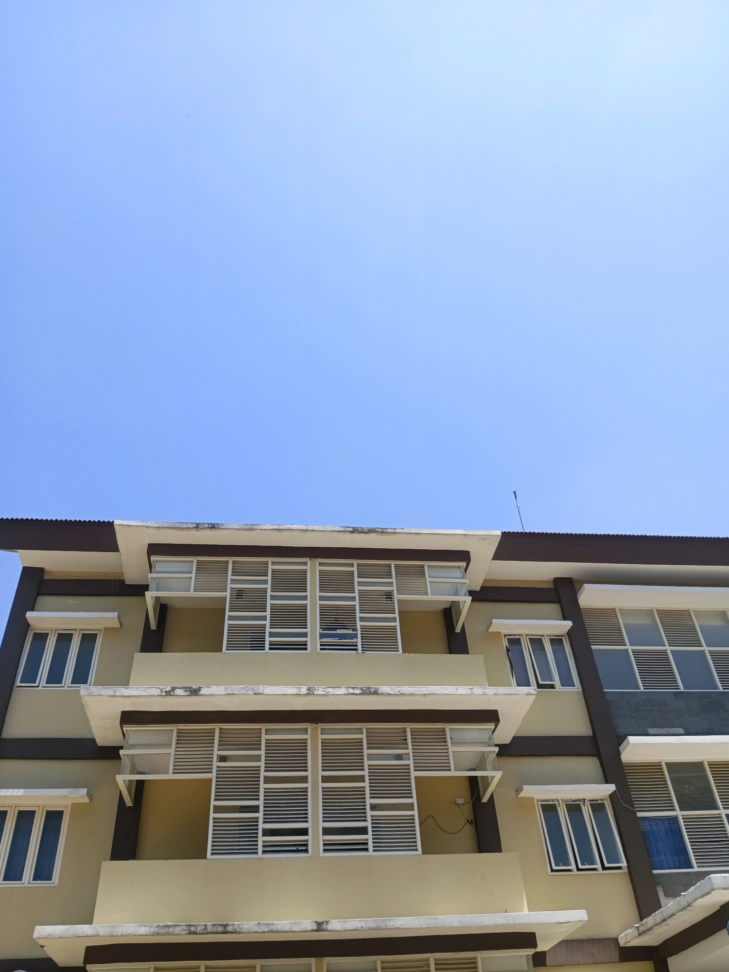 The image is a low-angle shot of a modern building with a clear blue sky above. The building appears to be multi-story, with distinct architectural features such as protruding balconies and numerous windows with white louvers. The facade is painted in muted tones of yellow and gray, with dark brown trim. The sky is a solid, bright blue, devoid of clouds, suggesting a sunny day. A thin, metallic antenna is visible on the roofline. The scene is static and devoid of human presence or activity. The overall impression is one of architectural documentation or a cityscape captured on a clear day.