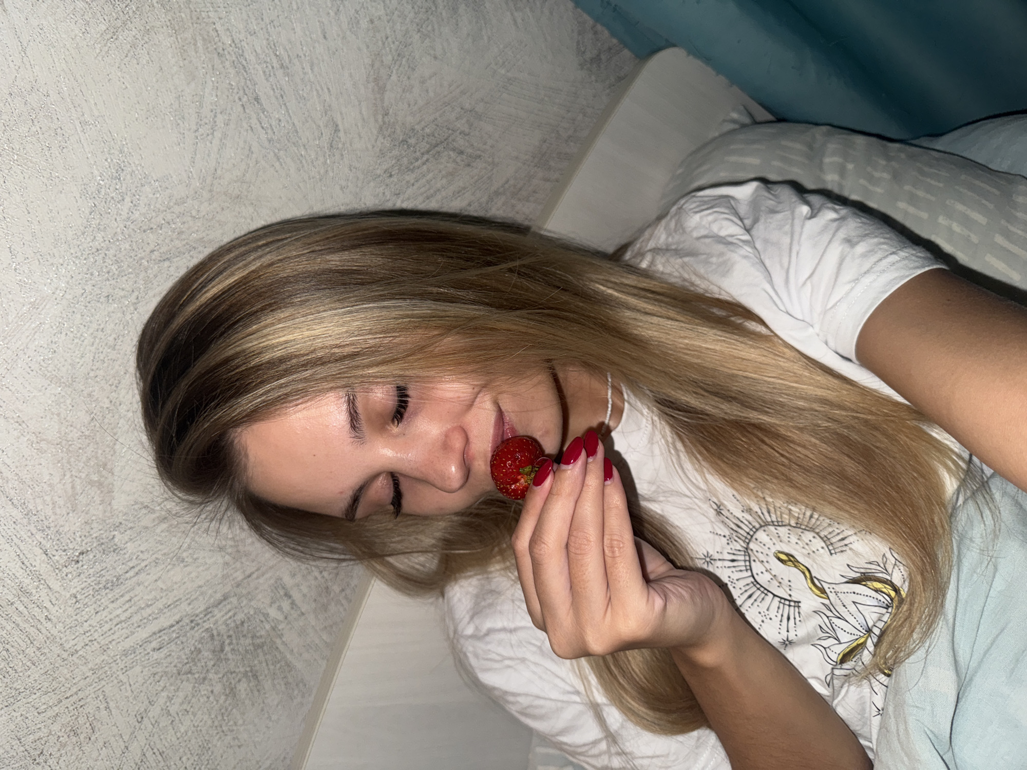 In Chaikovsky, Russia, a young woman with long blonde hair, wearing a white t-shirt with a circular design featuring a snake and celestial elements, is lying down. She is holding a single strawberry near her nose, with her eyes closed. Her fingernails are painted red. The background consists of a textured, light-colored ceiling and a partially visible turquoise curtain. The lighting suggests an indoor setting, possibly a bedroom, with a soft, ambient glow.