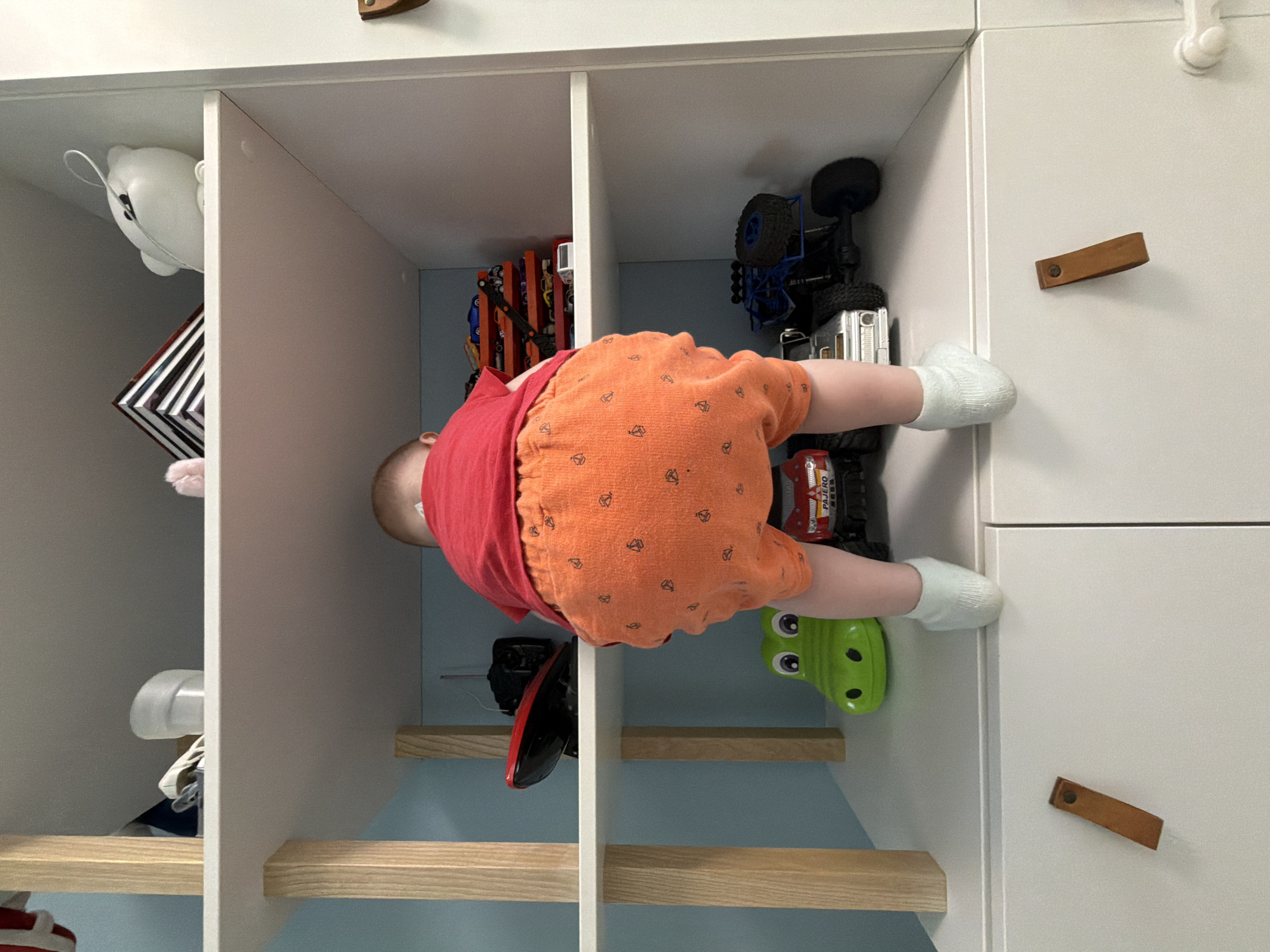 A baby wearing a red shirt, orange shorts, and white socks is lying on their stomach inside a white shelving unit filled with toys. The shelving unit is made of white painted boards and has wooden dowel supports. The toys visible include toy cars and a toy alligator. The shelving unit is situated against a pale blue wall. The location is unknown and there is no information to confirm that the image was taken in Votkinsk, Russia.
