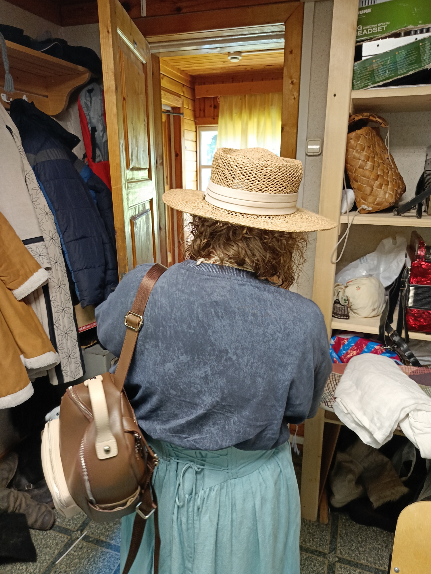 A person is standing in a room with wooden walls and a wooden door. The person is wearing a straw hat, a blue shirt, and a light blue skirt. They have a brown bag slung over their shoulder. The room appears to be a storage area, with shelves filled with various items, including coats, baskets, and fabrics. The environment suggests a rustic or rural setting, possibly a countryside home or a museum exhibit in Semenkovo, Russia, given the context. The lighting is dim, with natural light coming from a window visible through the open doorway.