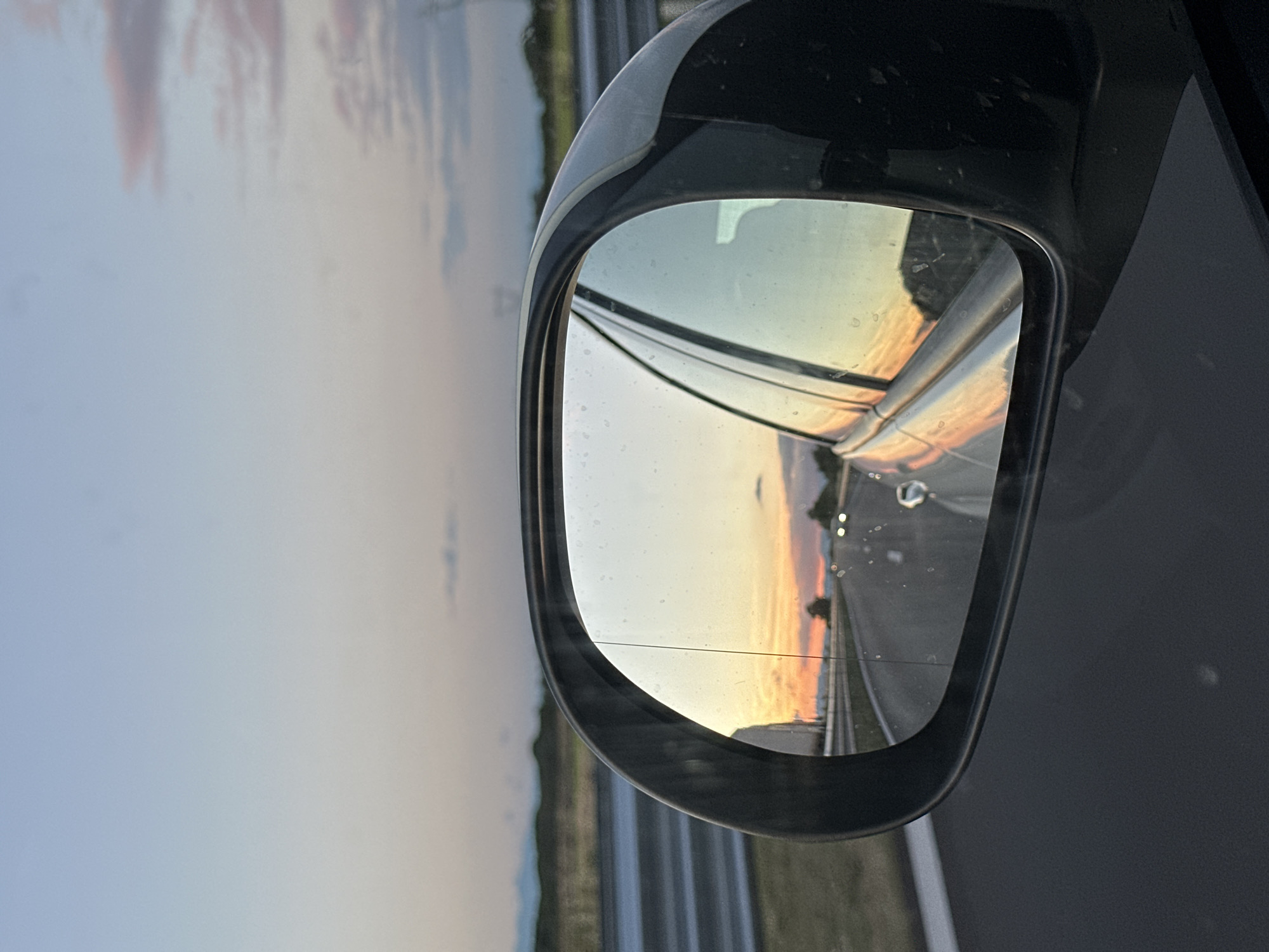 A side-view mirror of a vehicle reflects a sunset over a road. The sky in the reflection is a gradient of pale blue, pink, and orange, with faint clouds. The road ahead in the reflection appears to be an empty highway with a few car headlights visible in the distance. The exterior of the side-view mirror is black. The foreground of the image shows the dark gray car body. In the background outside the mirror, a guardrail runs along the side of the road, and a distant landscape of rolling hills and faint mountain ranges is visible under a pale sky. The location is the city of Malo Konare, Bulgaria.