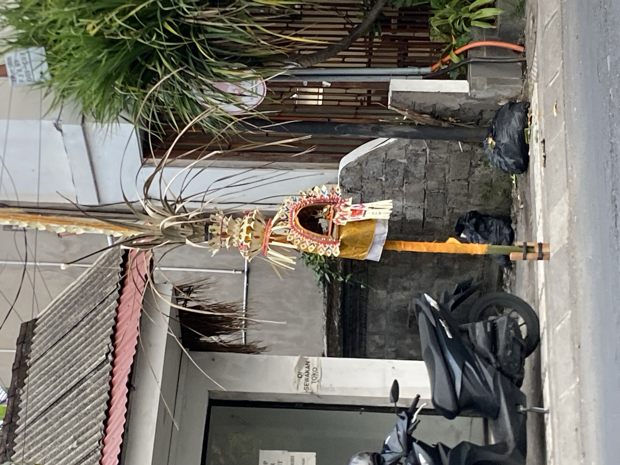 The image captures a street scene in Mengwi, Indonesia, with a focus on a decorative item placed on the sidewalk next to a building. The decorative item is tall and slender, made of dried palm leaves and adorned with colorful fabric and ornaments, suggesting a cultural or religious significance. It is supported by a yellow pole tied with dark bands at the bottom. To the left of this decoration, a black motorcycle is parked on the sidewalk. The building behind the decoration appears to be a shop or residence, with a grey corrugated metal roof and a white facade. A sign reading "DI SEWAKAN TOKO" (shop for rent) is visible on the wall. The environment shows signs of urban life, with a paved road in the foreground and scattered debris, including black trash bags, on the sidewalk. Lush green foliage and a tall plant with long leaves are visible on the right side of the image, partially obscuring a fence. The overall scene depicts an ordinary day on a street in Mengwi.