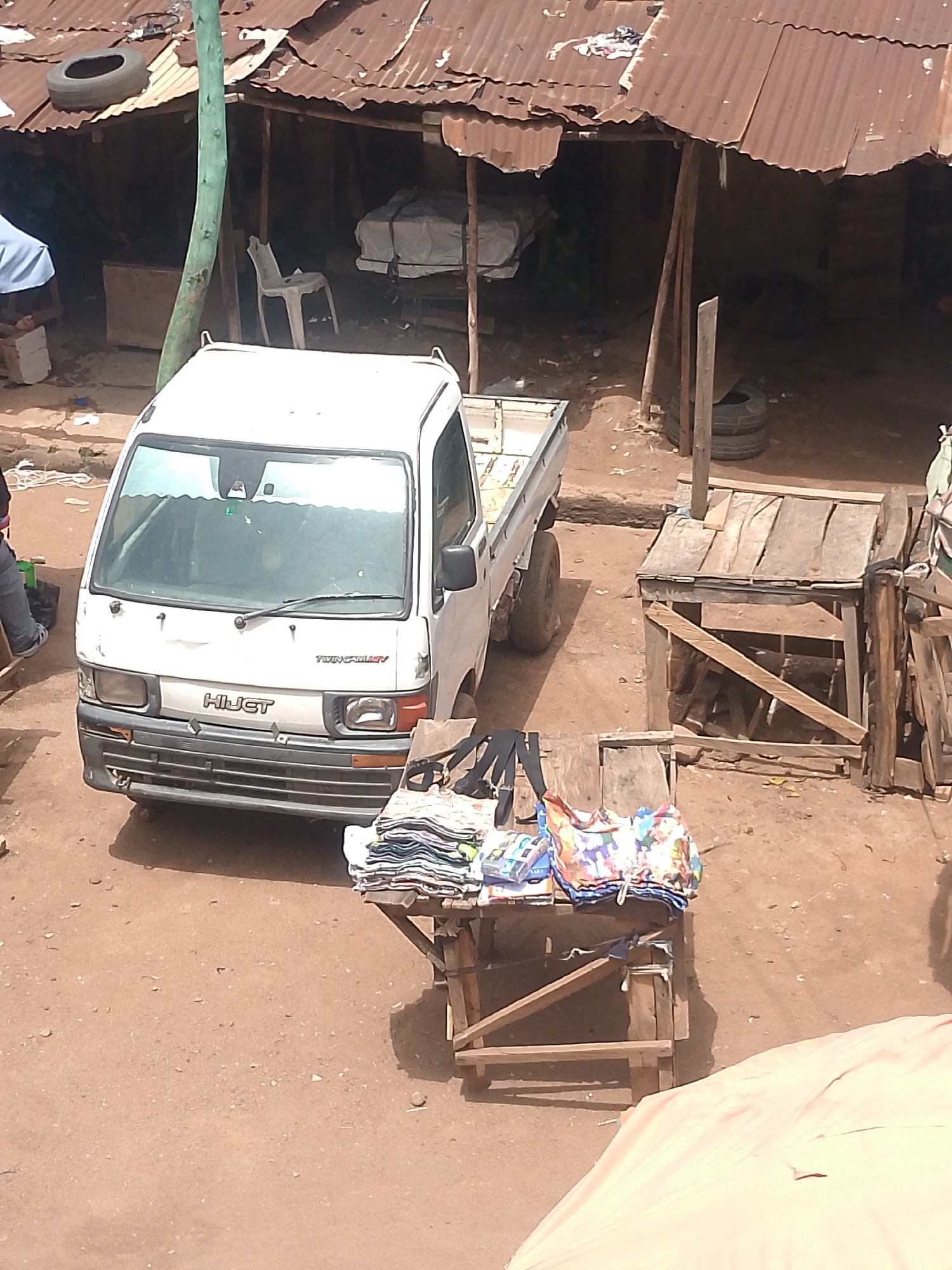 The image depicts a street scene in Bukuru, Nigeria. A white Daihatsu Hijet mini-truck is parked on a dirt road. In front of the truck, there is a wooden table with clothes and bags displayed for sale. Adjacent to the truck is a wooden stall. Overhead, a corrugated iron roof structure provides shade. To the left of the truck, an individual is visible, and several tires and a white chair are located under the shelter.