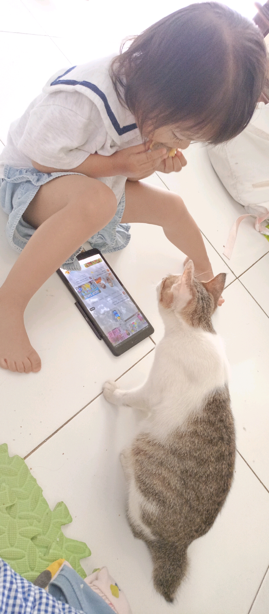 In a residential setting, likely a home floor in Kotabumi, Indonesia, a young child with dark hair, wearing a white shirt with a blue sailor collar and a light blue denim skirt, sits on the floor. The child is holding and eating a small yellow food item. In front of the child, a tablet is propped up, displaying a colorful interface that appears to be a video or game with cartoon characters. To the right of the tablet, a calico cat with patches of white, brown, and grey fur lies on its side, looking up at the tablet. The floor is tiled in a light color, and a green patterned play mat and some clothing items are visible in the foreground. The scene suggests a moment of shared attention between the child and the cat towards the tablet's content.