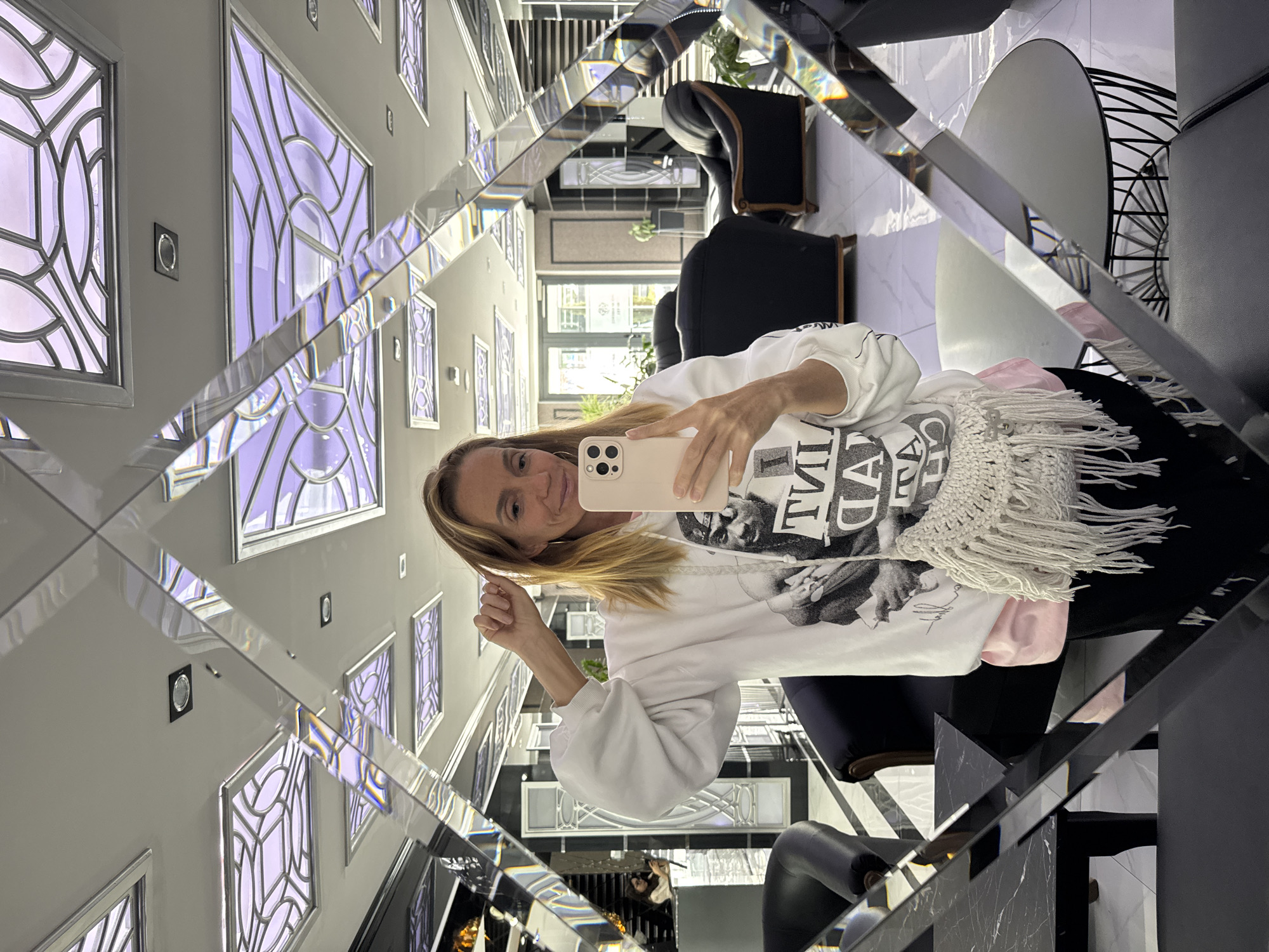 A selfie taken in Tashkent, Uzbekistan, shows a woman with blonde hair, wearing a white sweatshirt, holding a smartphone, presumably taking a picture of herself in a diamond-shaped mirror. The mirror reflects a hallway with decorative window panels with purple lighting, black chairs, and a round table. The background reveals a glimpse of an outdoor area.