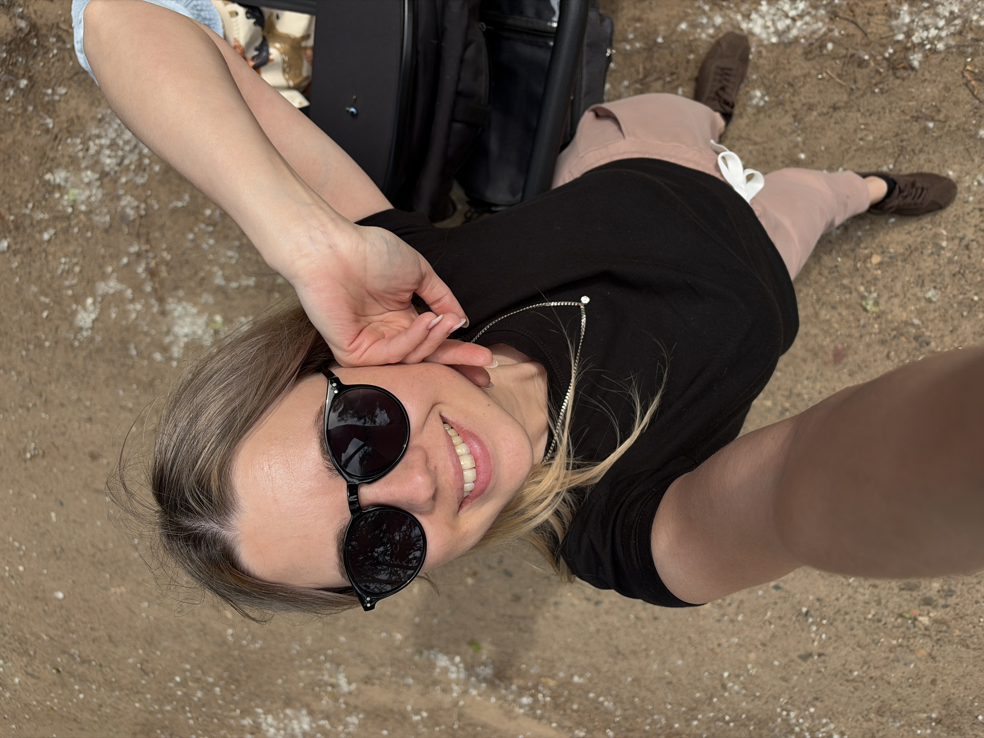 The image presents a young woman captured from a high-angle, top-down perspective, appearing to be lying on the ground while taking a selfie. She has light brown hair, parted slightly, and is wearing black, round-framed sunglasses. Her face is turned upwards, directly at the camera, with a wide, joyful smile revealing her teeth.
She is dressed in a black short-sleeved t-shirt, which features a silver chain with a small white pendant or decorative zipper detail near the neckline. Her lower body is clad in light brown or pink pants with visible white drawstrings tied in a bow. She wears brown lace-up shoes.
Behind her head and upper torso, parts of black luggage, including what appears to be a suitcase and another bag, are visible. The scene is outdoors, with the ground composed of sandy or fine dirt material, liberally scattered with small white pebbles or aggregate. The overall lighting suggests it is daytime. The woman's broad smile conveys an emotion of happiness, contentment, or playfulness. There is no visible text in the image.