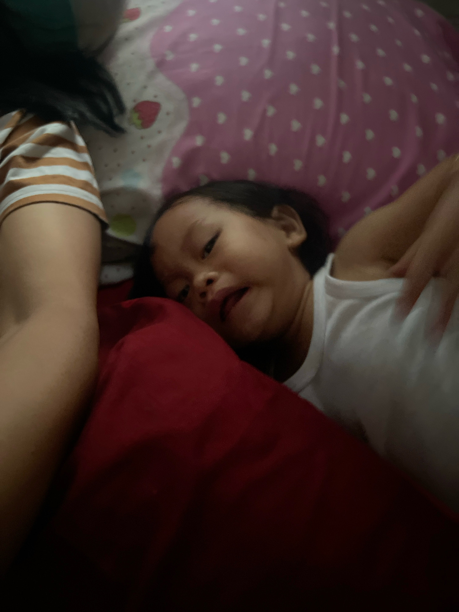 In the dimly lit interior of a home in Kecamatan Pati, Indonesia, a young child, likely of Southeast Asian descent, lies on their side on a red pillow. The child, wearing a white tank top, has dark hair and is looking towards the viewer with a slightly distorted facial expression. To the left of the child, an adult's arm, clad in a horizontally striped t-shirt (tan and white), is visible. The background consists of a pink pillow with a pattern of white hearts and a strawberry graphic. The overall scene suggests a casual, intimate moment, possibly of the child being held or resting near an adult.