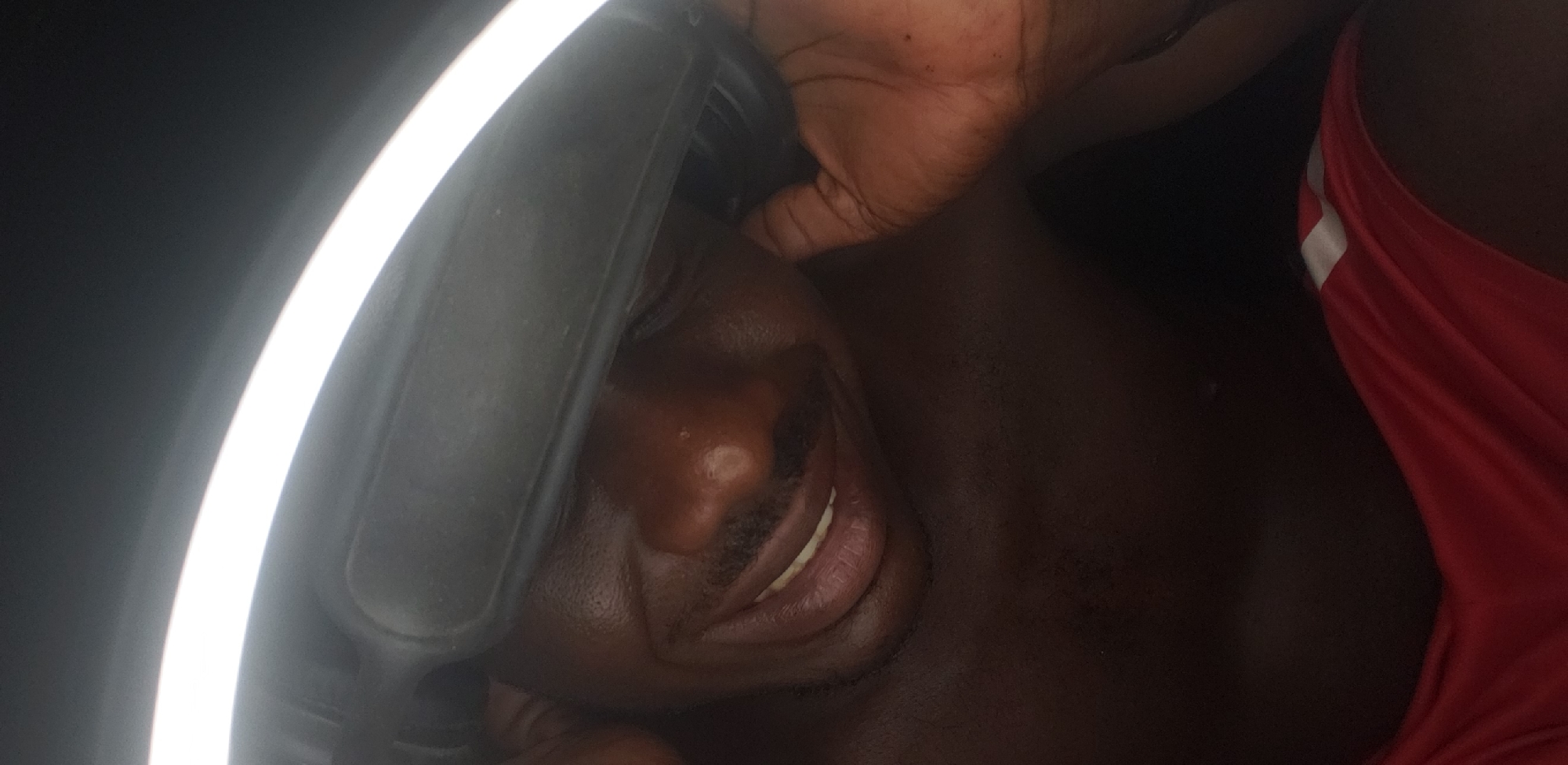 A dark-skinned man is lying down, illuminated by a ring light. He is wearing a red garment with white stripes. The location is Narayi, Nigeria.