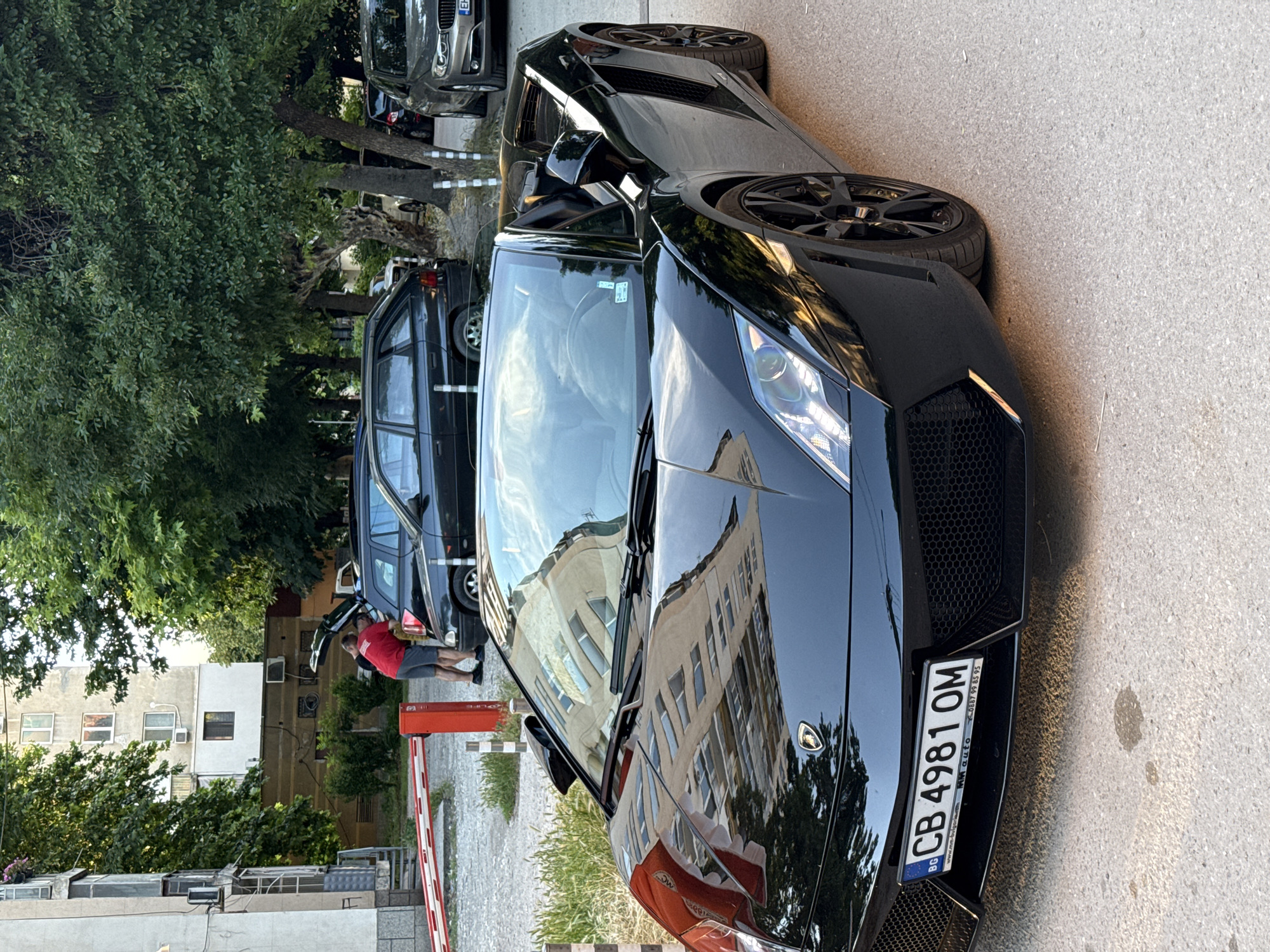The image shows a black convertible Lamborghini parked on a paved street in an urban environment. In the background, there are several cars parked, including a dark station wagon with its trunk open and two men near it. A red and white barrier is visible in the left part of the image, and apartment buildings and trees line the street. The car has a Bulgarian license plate that reads "CB 4981 OM".