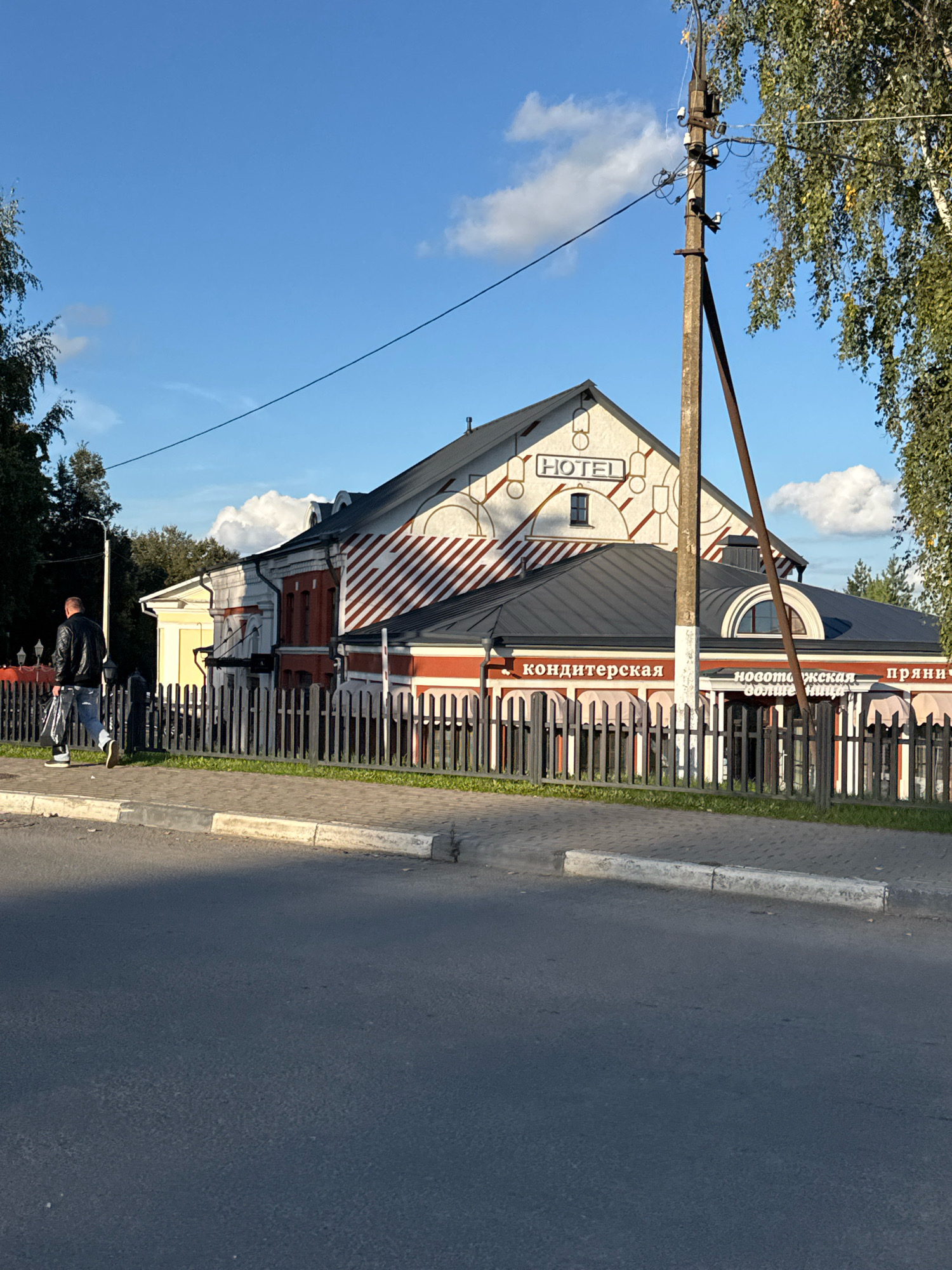 In Torzhok, Russia, a man wearing a black leather jacket and jeans walks away from the camera on a sidewalk next to a black metal fence. Beyond the fence is a building with red and white striped siding and a gray roof. The building has a sign that reads "HOTEL" in white letters. Below the hotel sign, on the lower part of the building, there are signs that read "КОНДИТЕРСКАЯ" (confectionery) and "новоторжская пряничная" (Novotorzhskaya Gingerbread House). A utility pole with wires stands in front of the building. The sky is blue with some white clouds. The ground in the foreground is asphalt.