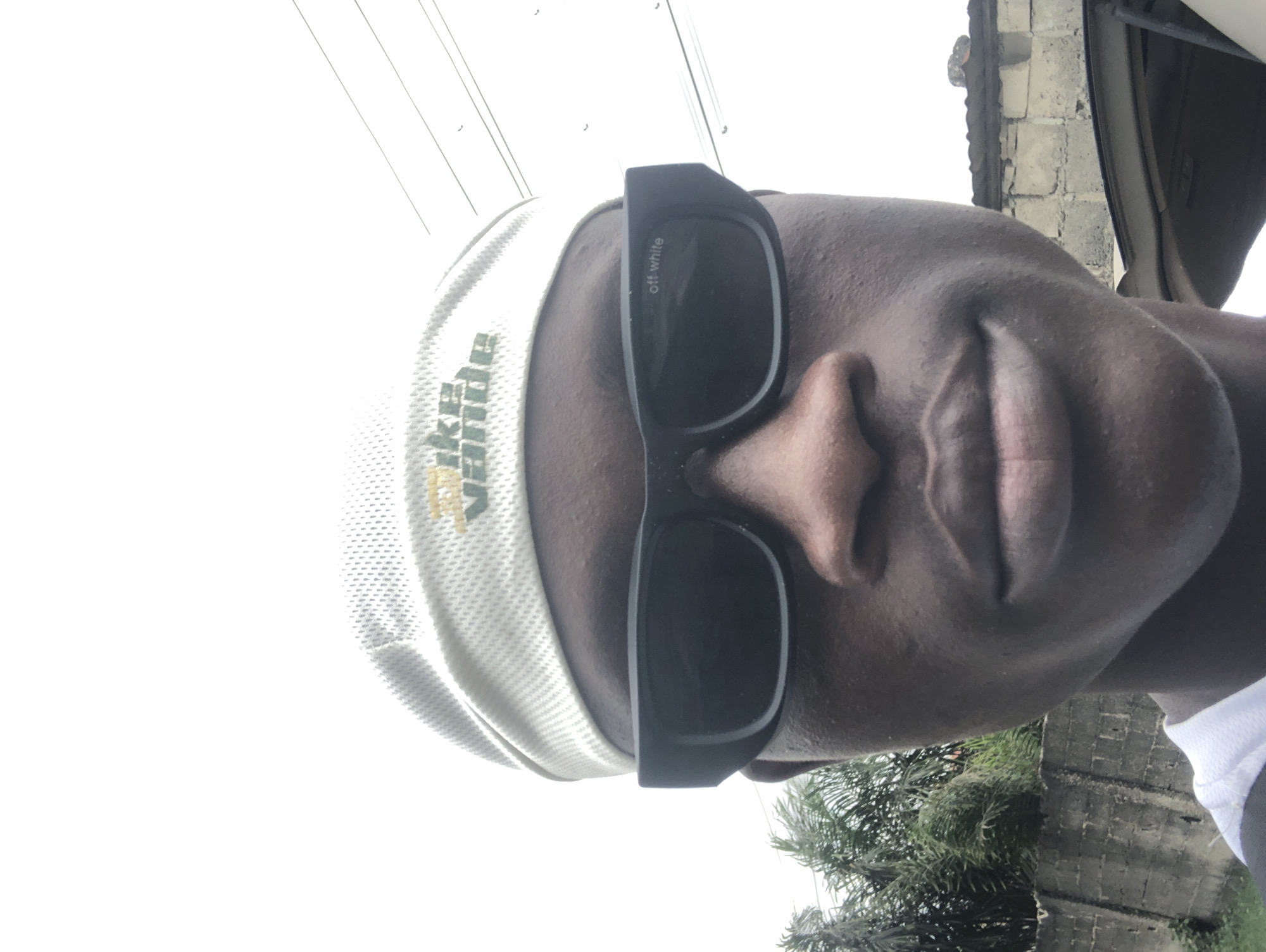 A selfie of a young Black male wearing a white and yellow bandana and black sunglasses. The bandana has the text "Nike Value" printed on it. The sunglasses have "off white" printed on the right lens. The background shows a brick wall, palm trees, and a clear sky with power lines. The image appears to have been taken outdoors in Ado, Nigeria.